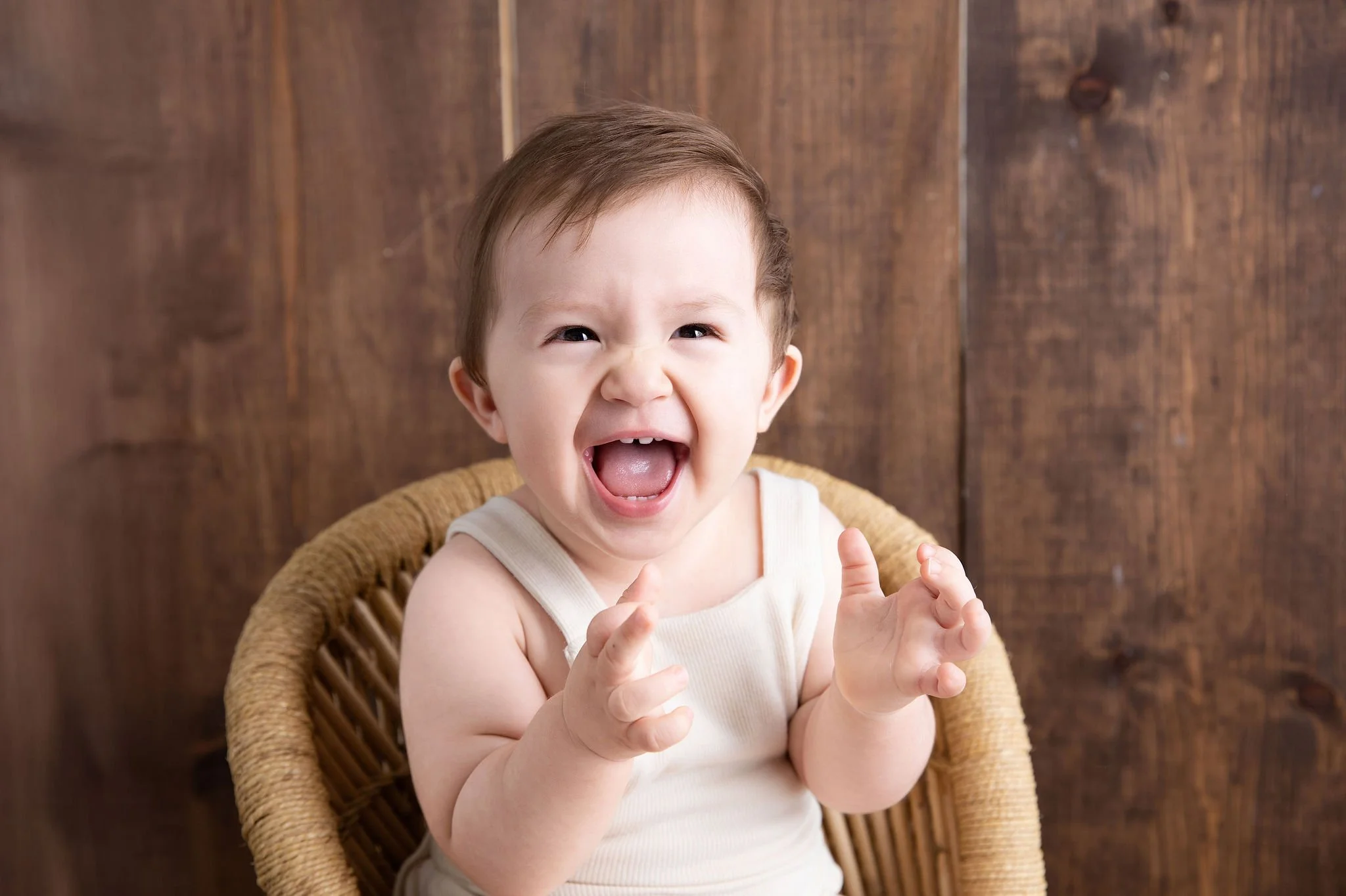 Young child with short brown hair, laughing or shouting, sitting in a woven rattan chair with a wooden wall in the background.