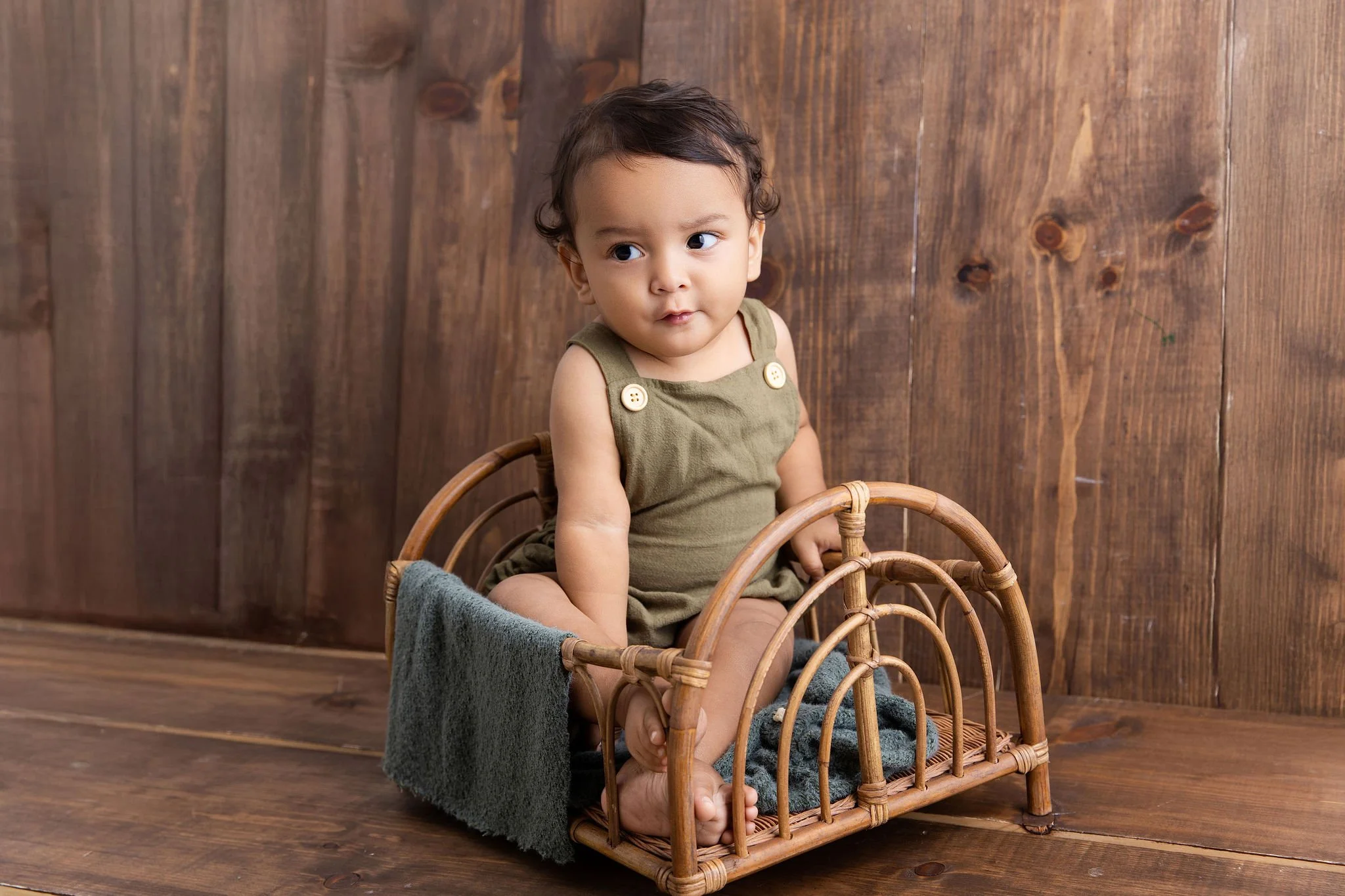 A young child sitting inside a small wicker basket, with a wooden wall background and a blanket draped over the side of the basket.