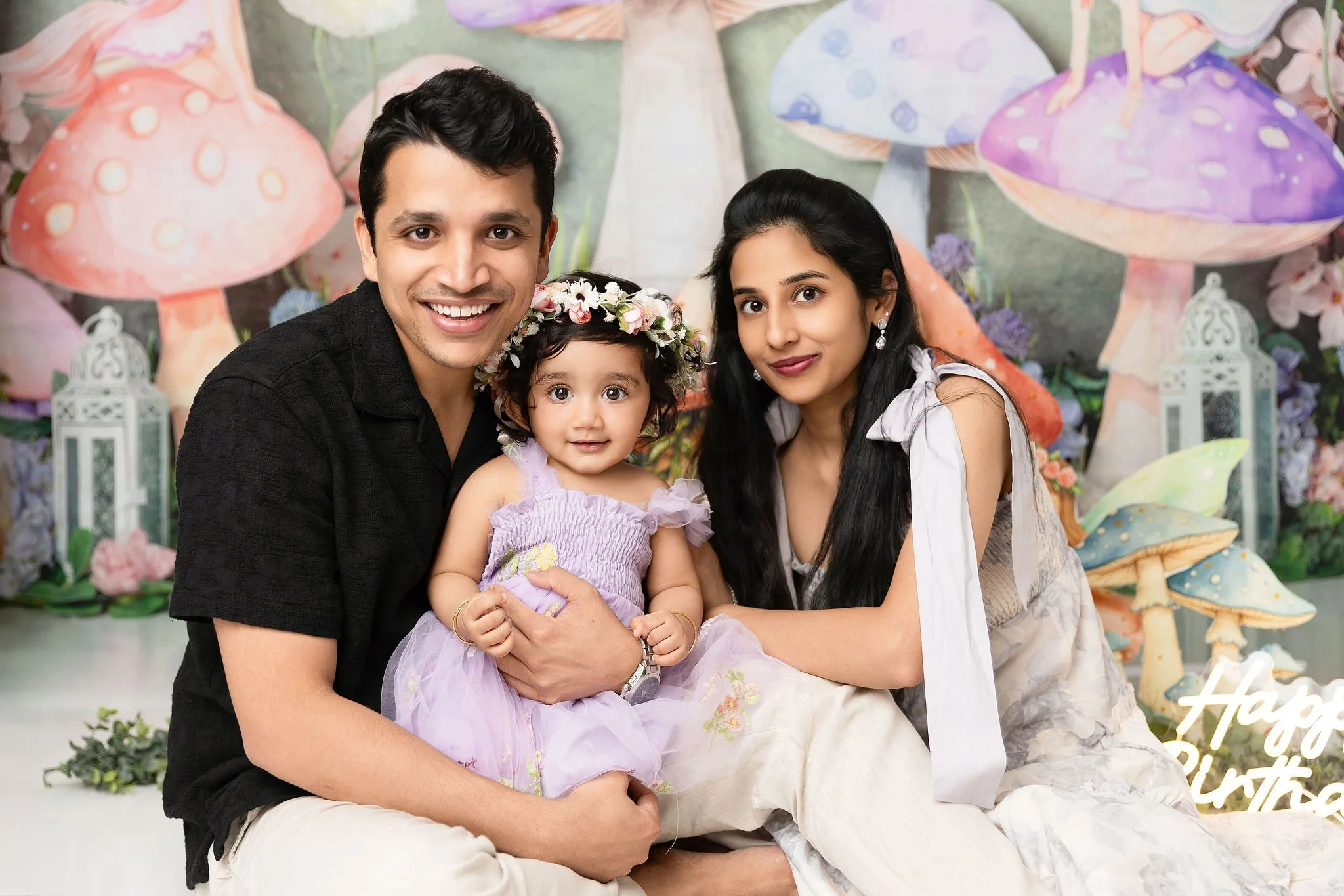 A happy family of three, including a man, a woman, and a young girl, sitting together at a birthday celebration with a colorful fairy tale backdrop featuring large mushrooms and lanterns.
