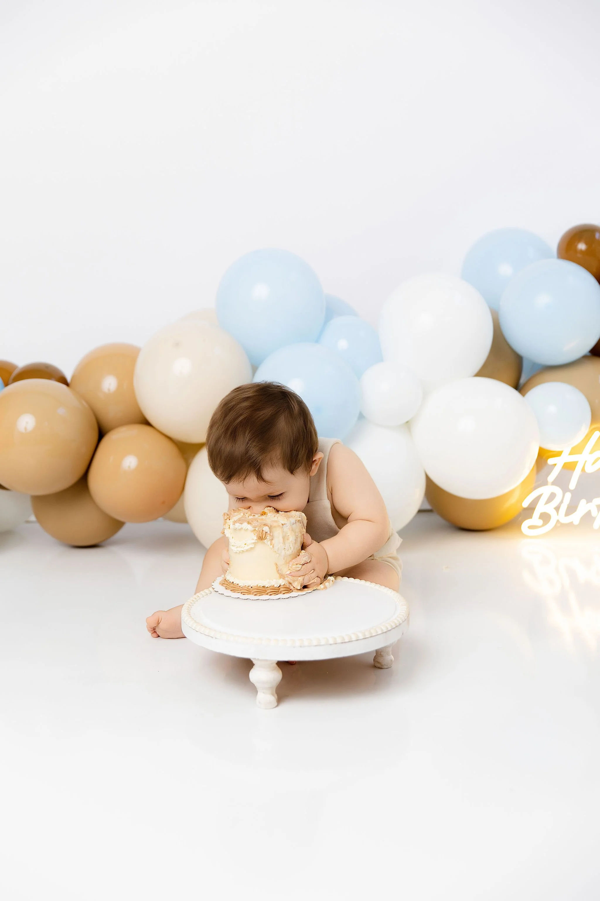 Young child in beige outfit biting into a small, partially eaten birthday cake on a white cake stand, with a backdrop of blue, white, tan, and brown balloons and a neon