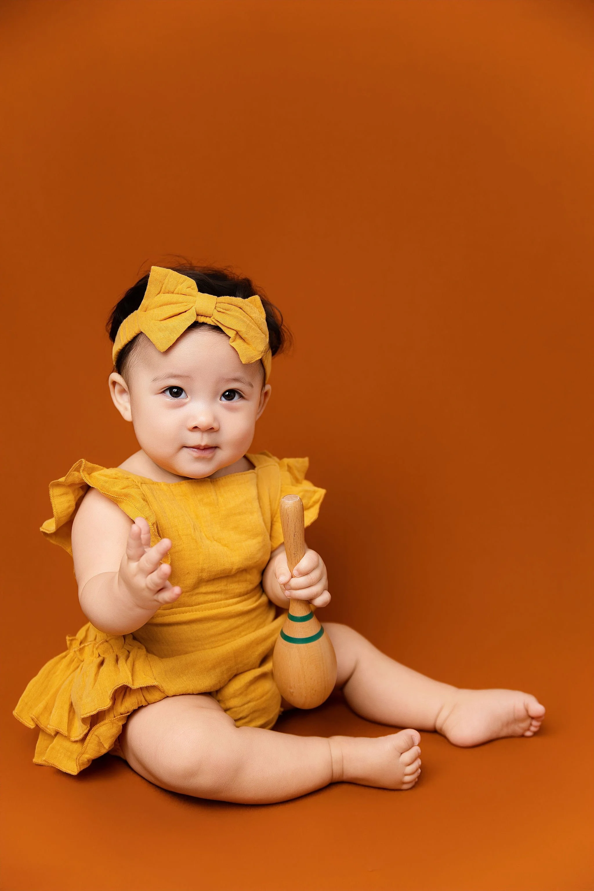 A baby girl with a yellow headband and dress holding a wooden maraca, sitting on a brown background.