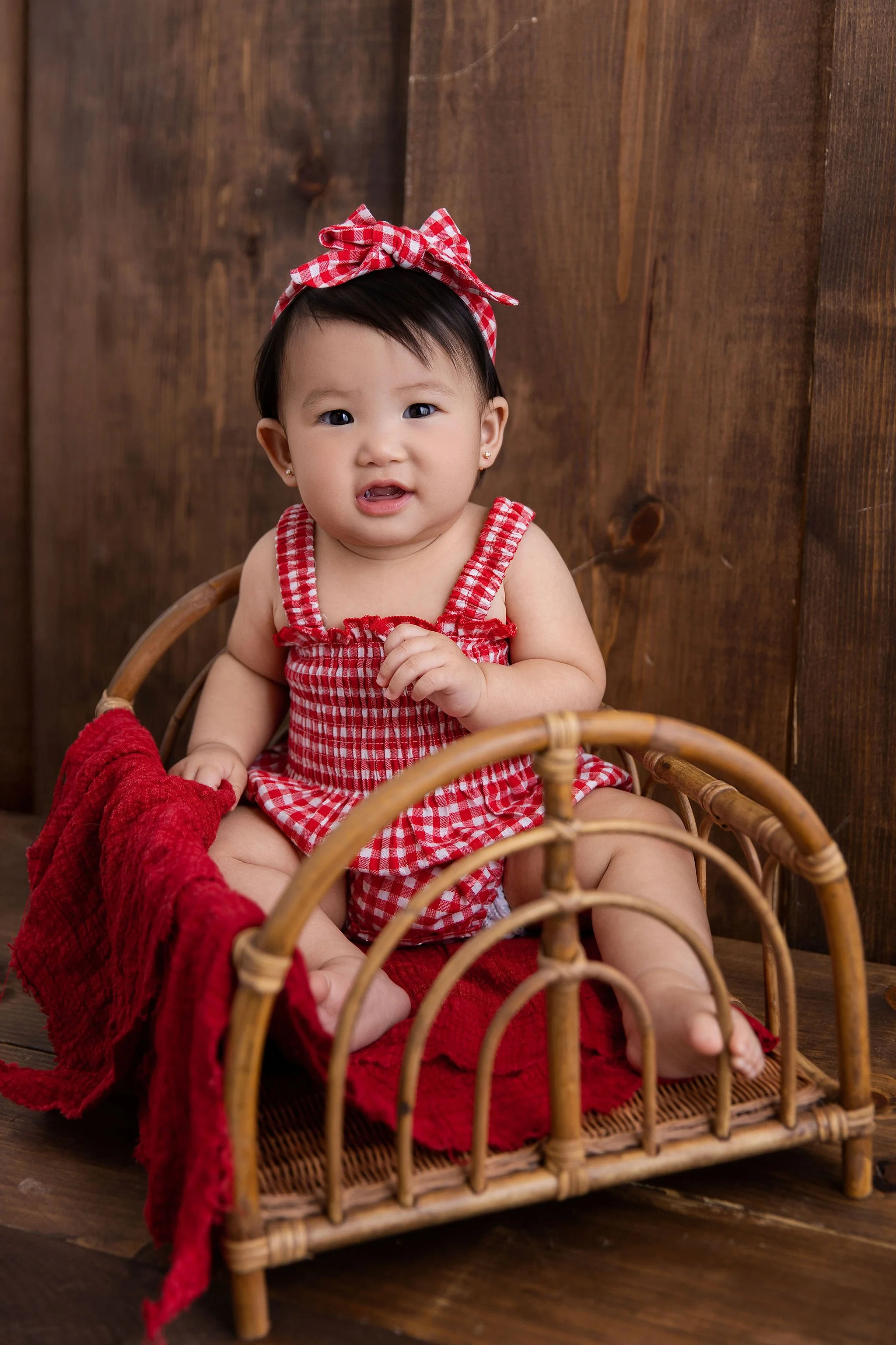 A baby girl wearing a red and white checkered dress and matching headband, sitting in a wicker bed with a red blanket against a wooden backdrop.