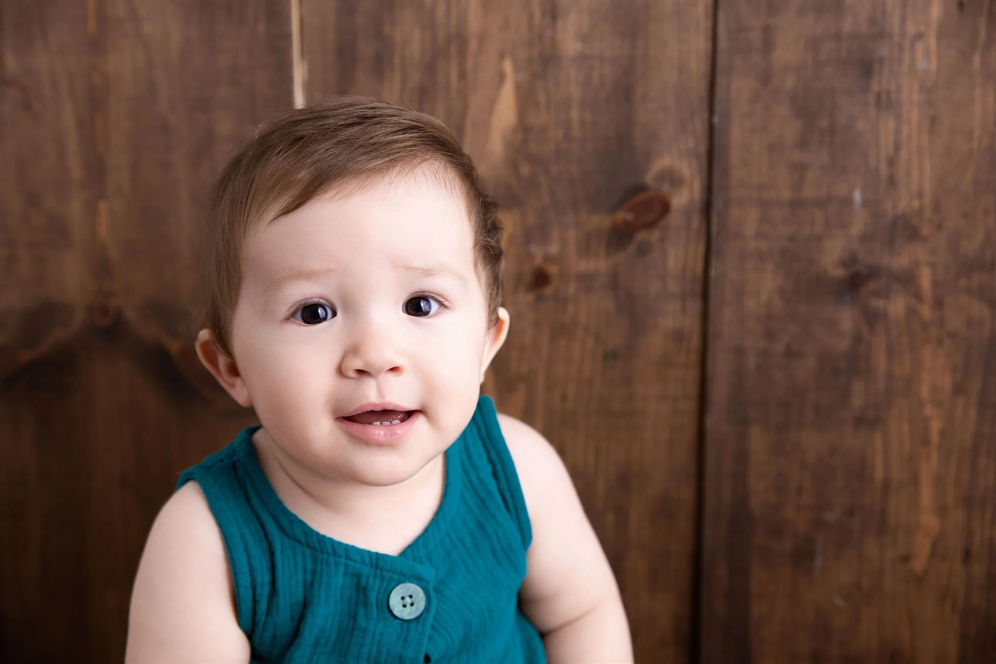 A young child with brown hair and big eyes looking at the camera, wearing a blue sleeveless shirt with a button, standing in front of a wooden background.