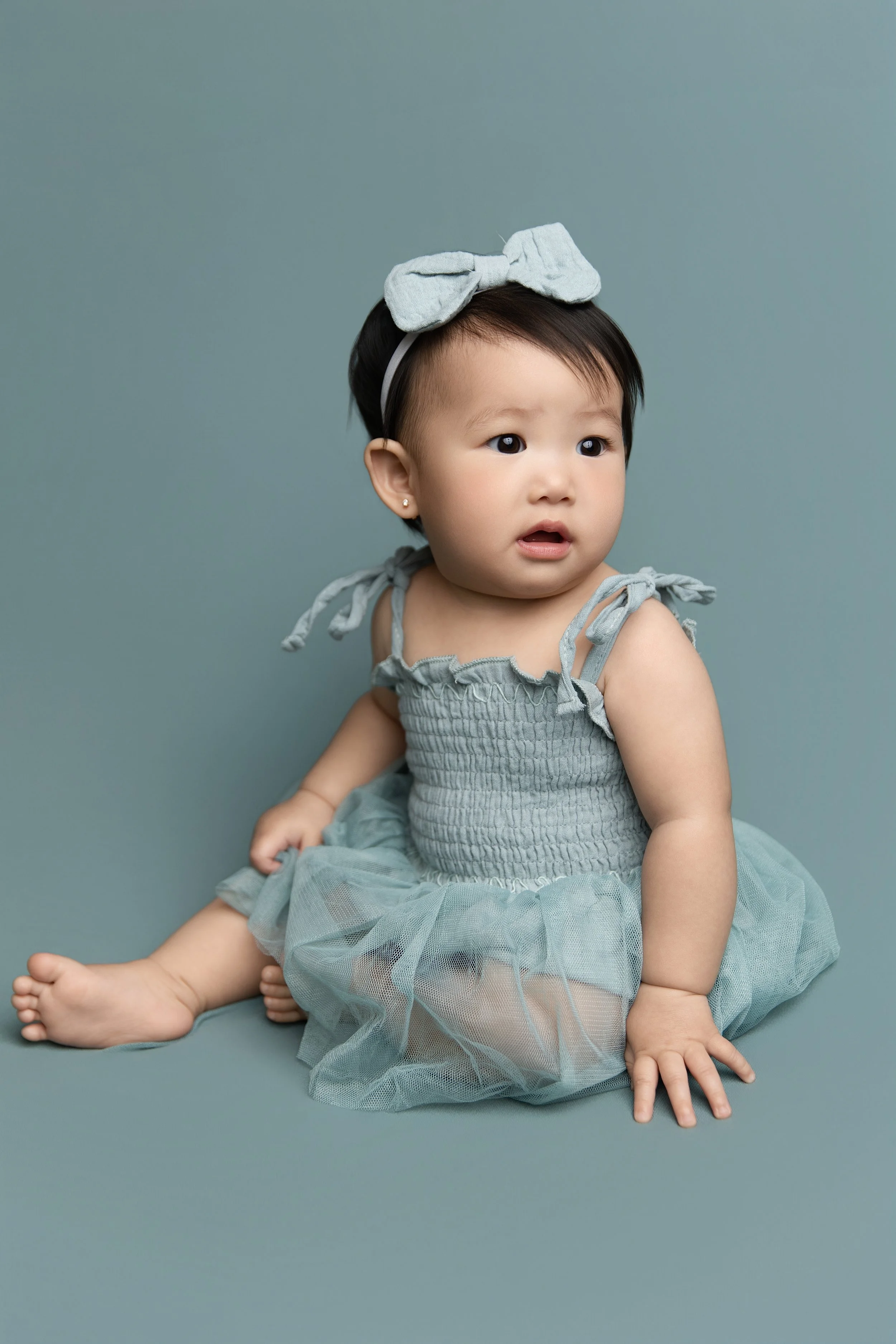 A young Asian girl wearing a gray dress with tied straps and a matching bow headband, sitting on a muted blue backdrop with a curious expression.