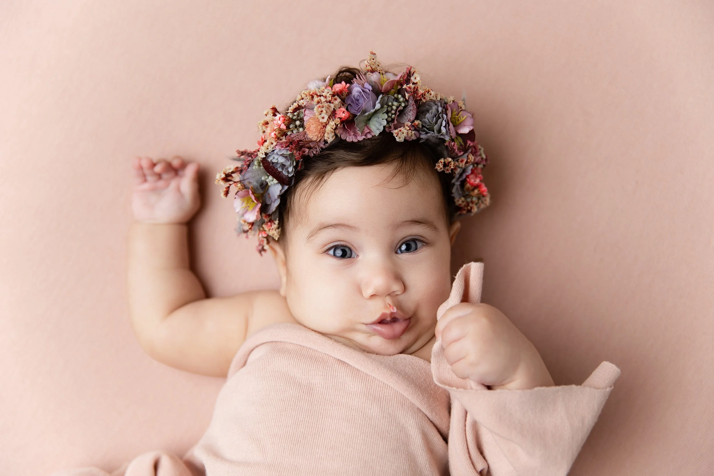 Adorable baby girl lying on a pink surface, wearing a floral crown and a pink dress, with bright blue eyes and chubby cheeks, holding one hand up.