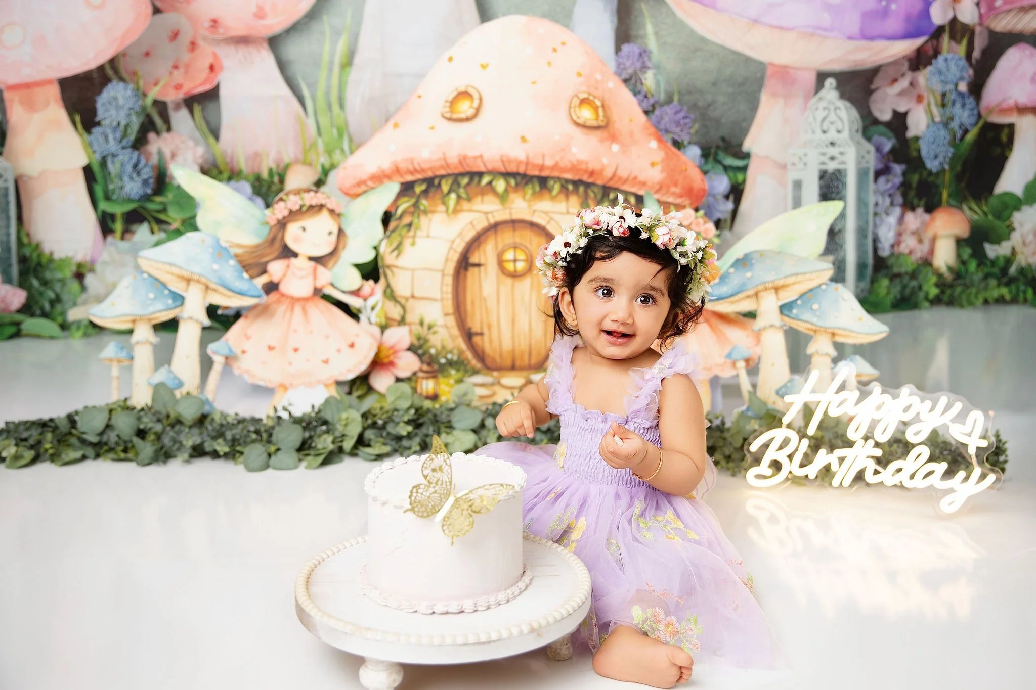 A young girl wearing a purple dress with a flower crown sits in front of a fairy tale-themed birthday backdrop. The backdrop includes whimsical mushroom houses, fairy characters, and lush flowers. There is a white birthday cake with butterfly decorat