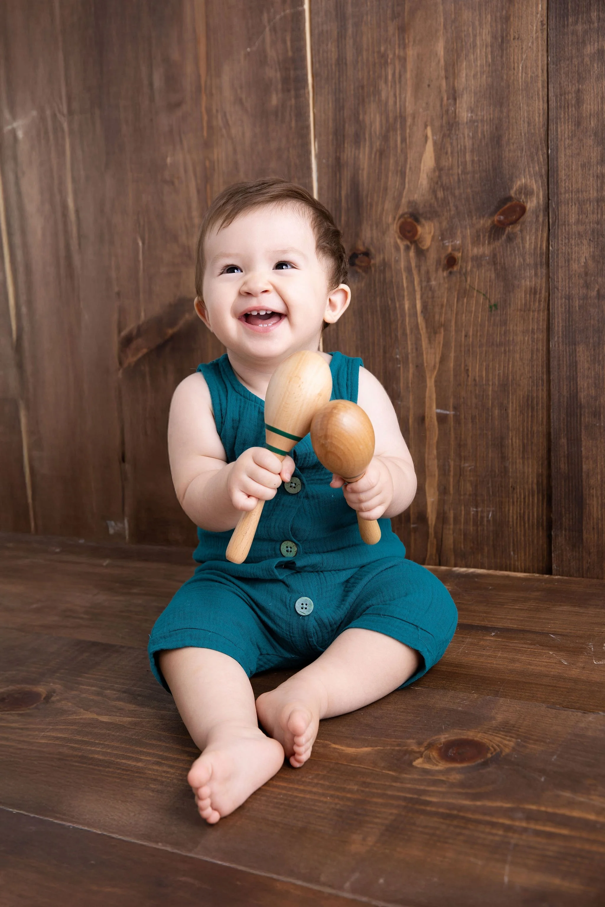 Smiling toddler sitting on a wooden floor holding two wooden maracas, wearing a teal sleeveless outfit with buttons.