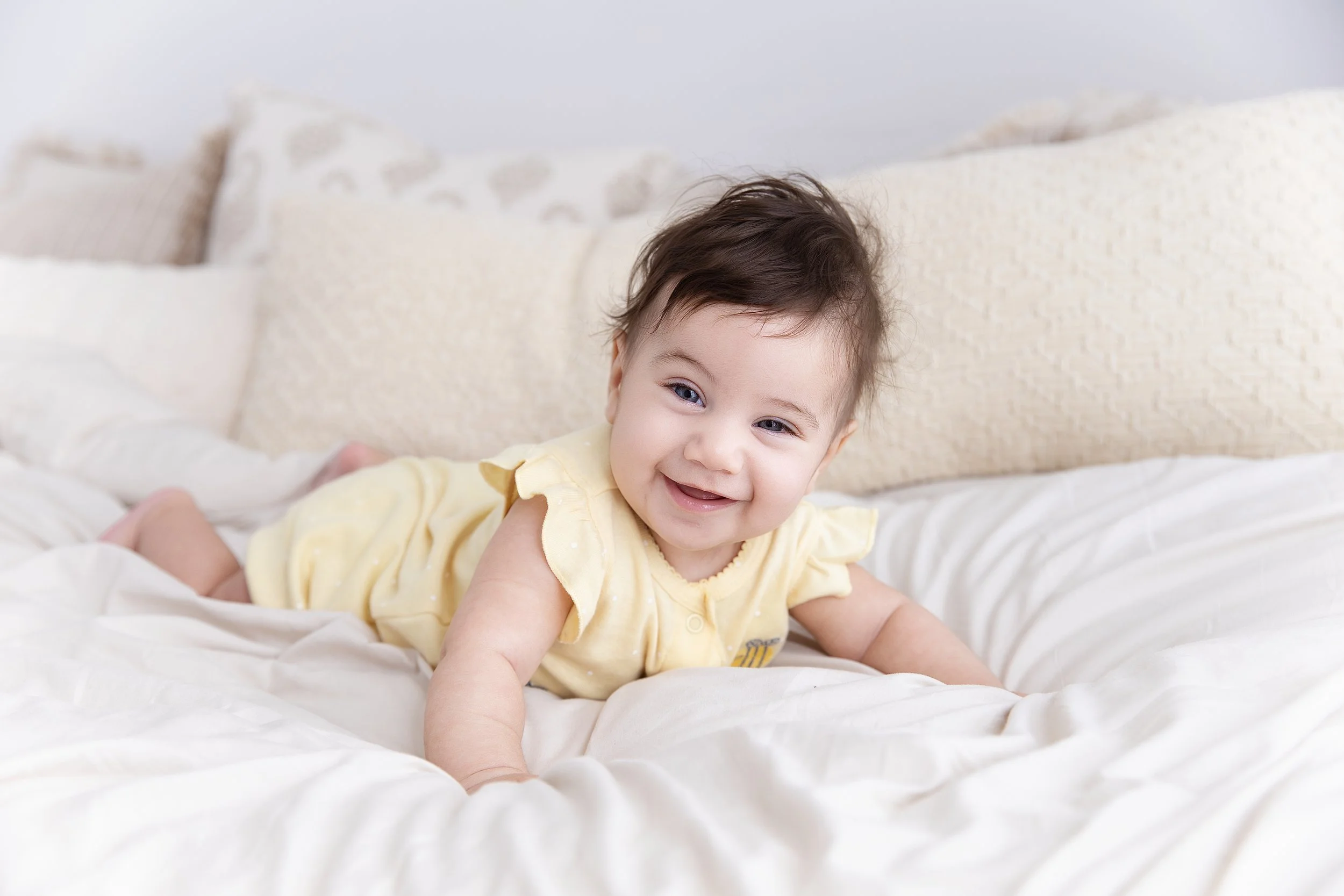 Smiling baby girl with dark hair in a yellow outfit, crawling on a white bed with cream-colored pillows in the background.