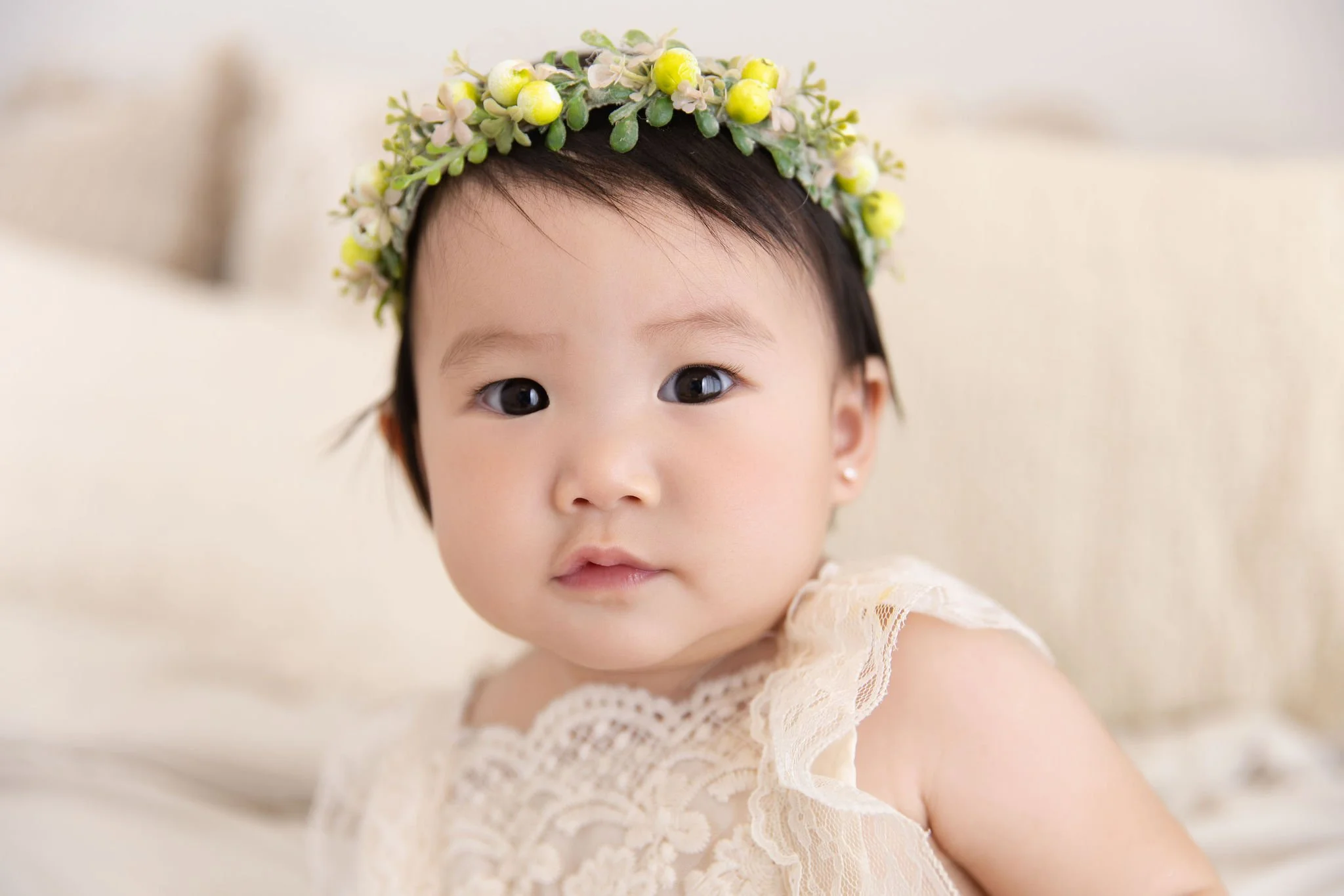 Young girl wearing a cream lace dress with a floral crown on her head, sitting indoors with a neutral background.
