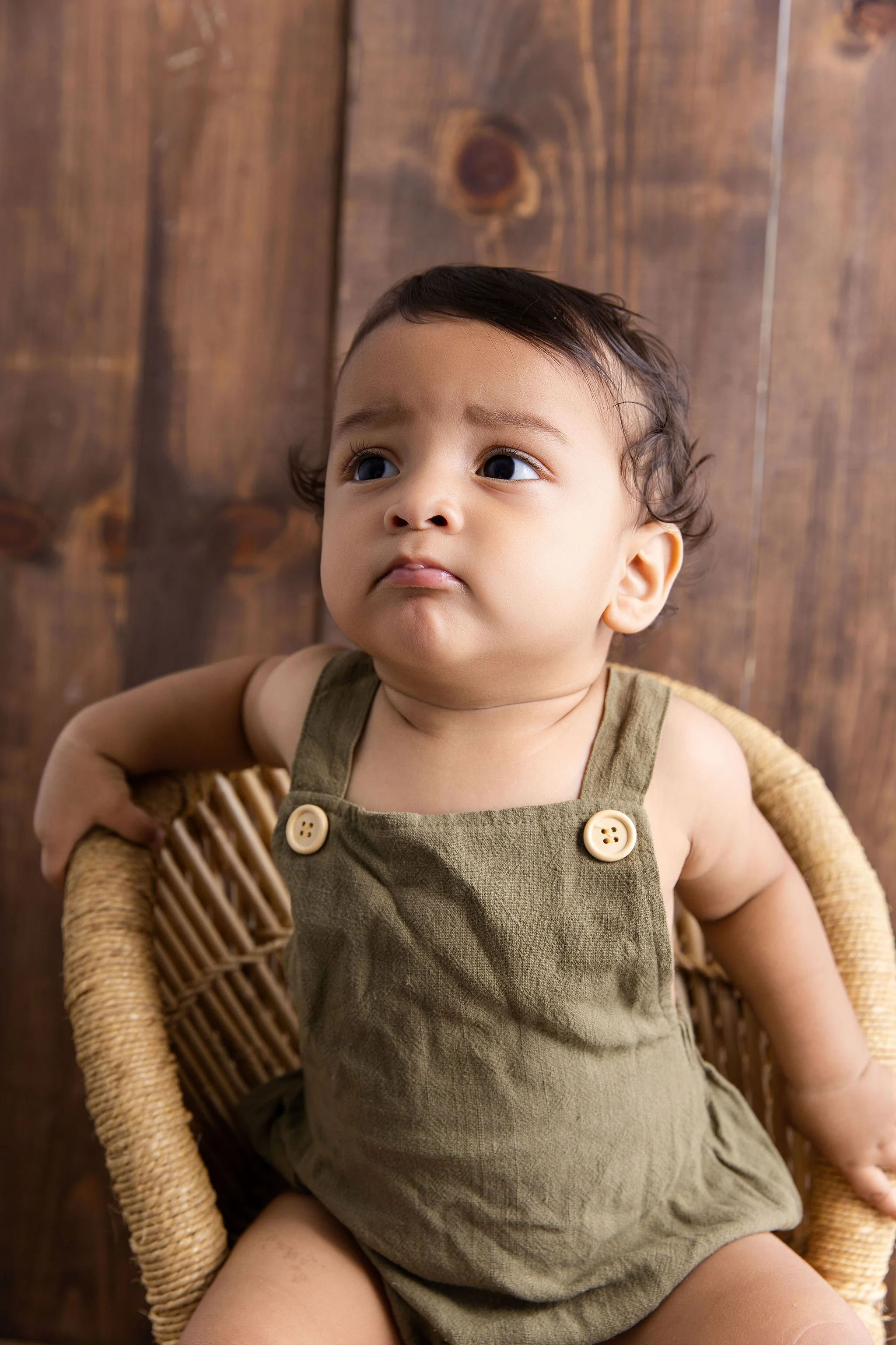 A baby sitting in a wicker chair with a wooden wall background, wearing a khaki romper with buttons, looking upward with a serious expression.