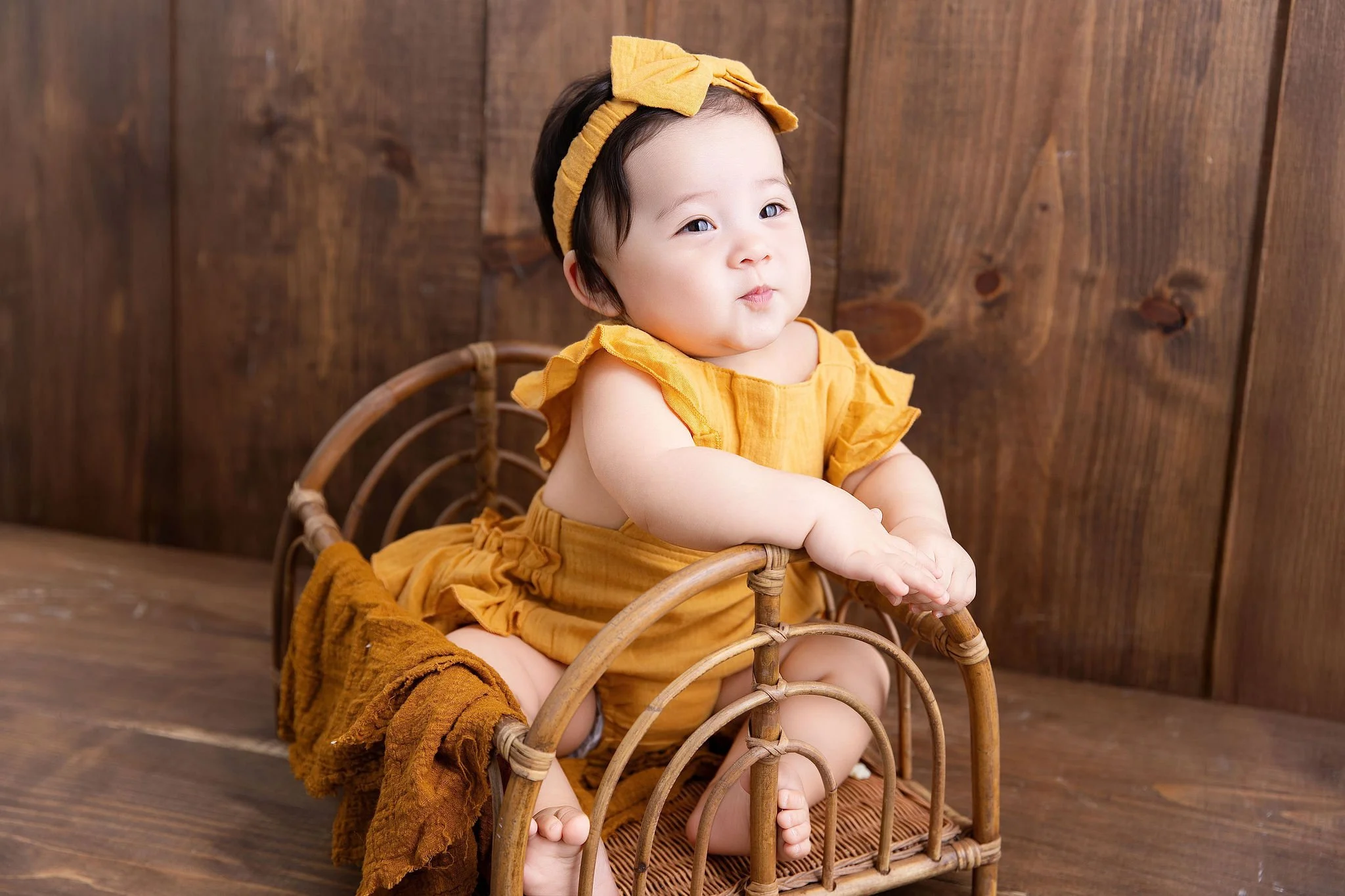 A baby girl sitting in a small woven wicker bed, wearing a yellow outfit with ruffle sleeves and a matching headband, against a wooden wall background.