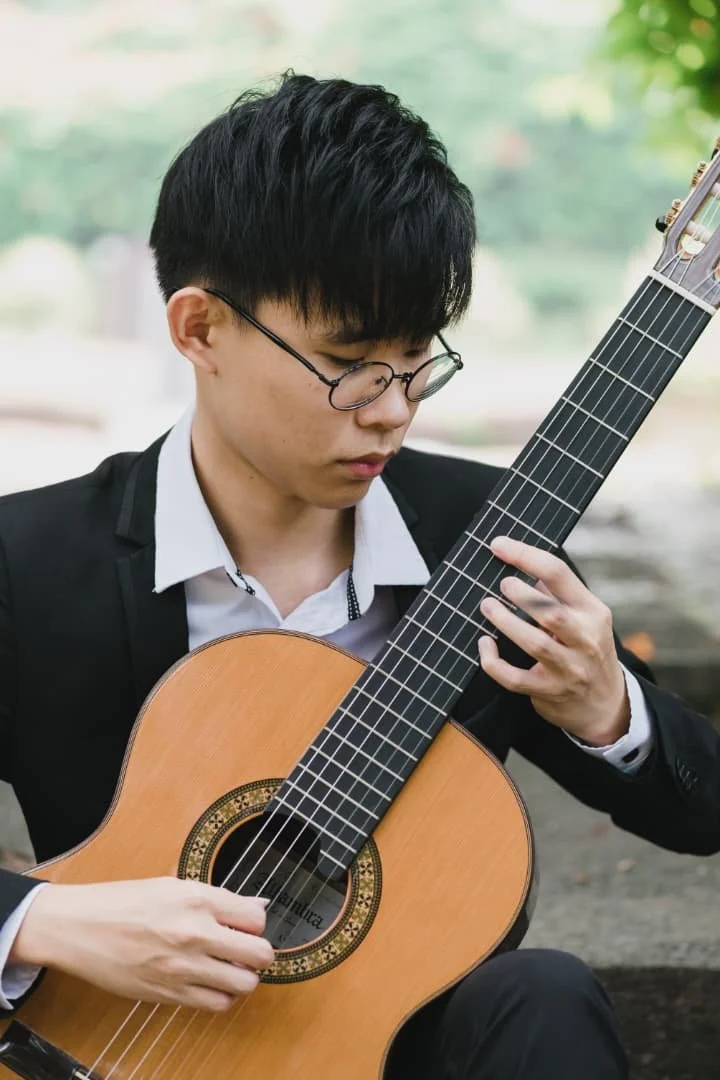 A young man with black hair and glasses wearing a black suit jacket and white shirt playing a classical guitar outdoors.