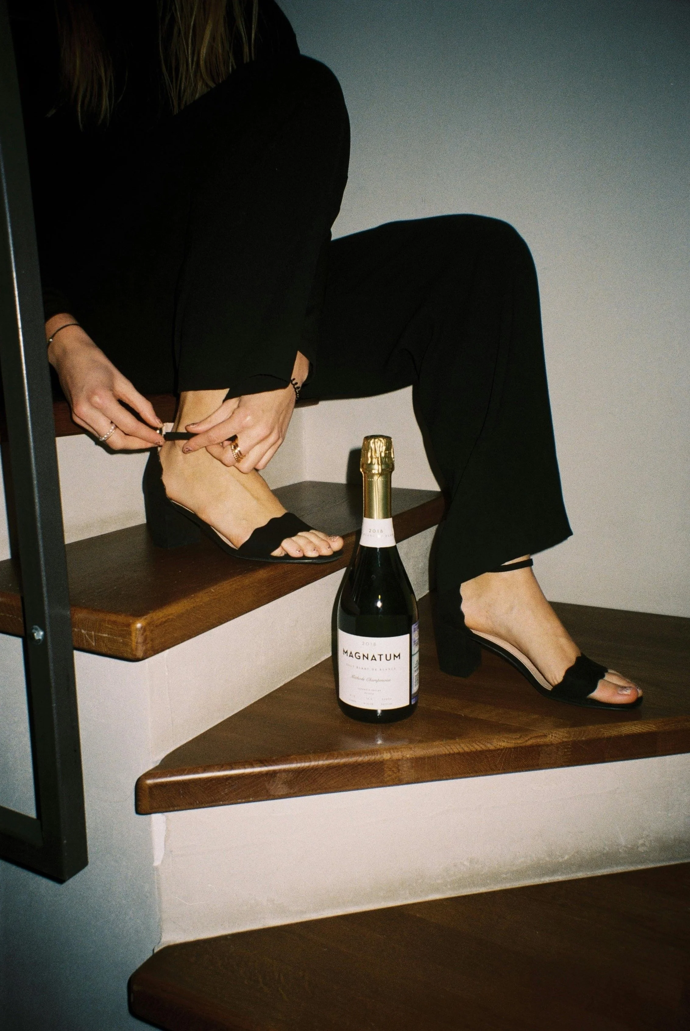A woman in black clothing is sitting on a staircase, moving a high-heeled sandal onto her foot, with a bottle of Magnum Champagne on a step below her.