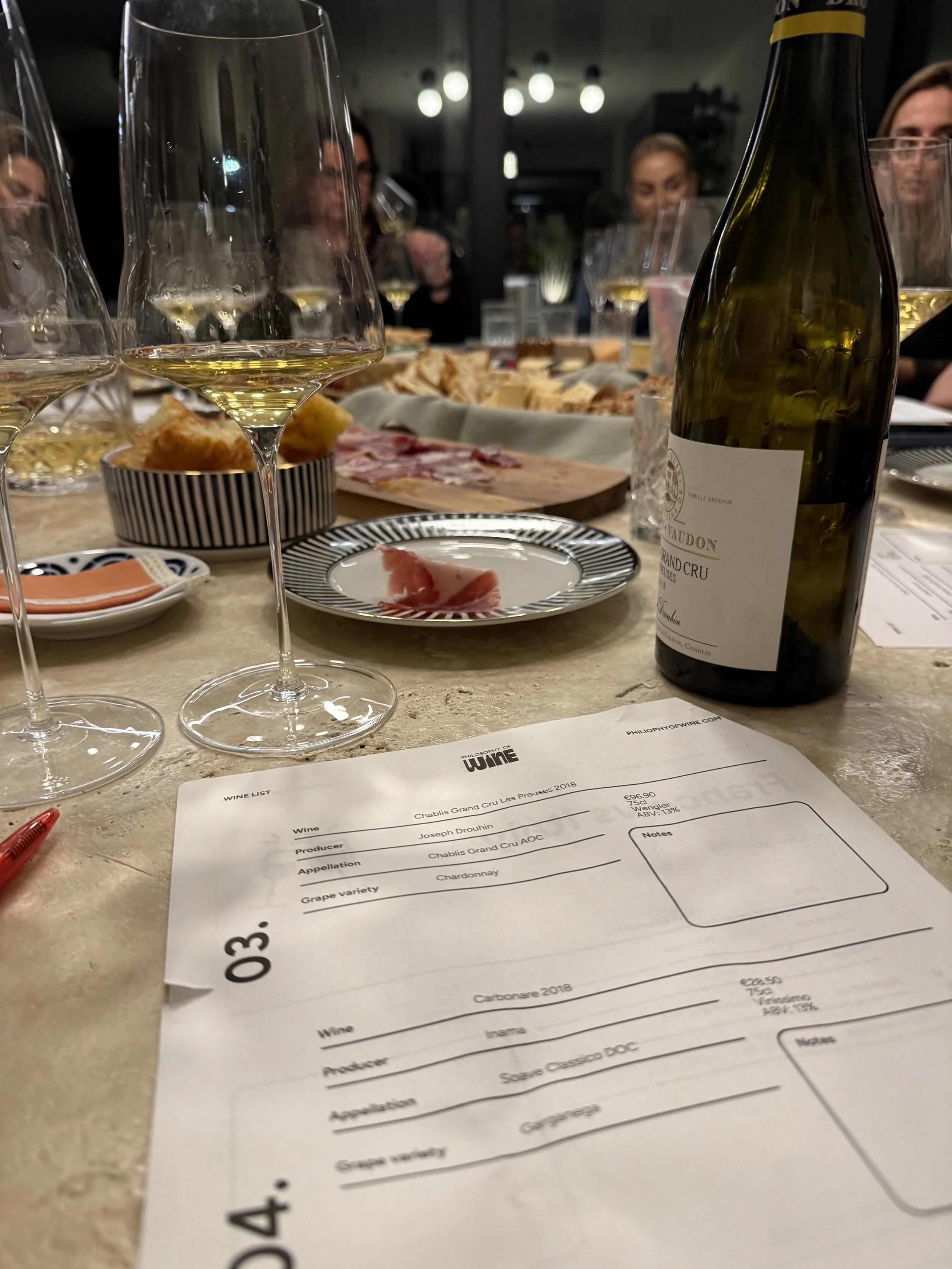 A table with glasses of white wine, a bottle of white wine, a plate with a slice of prosciutto, a tray with bread and charcuterie, a menu, and several people in the background enjoying a meal.