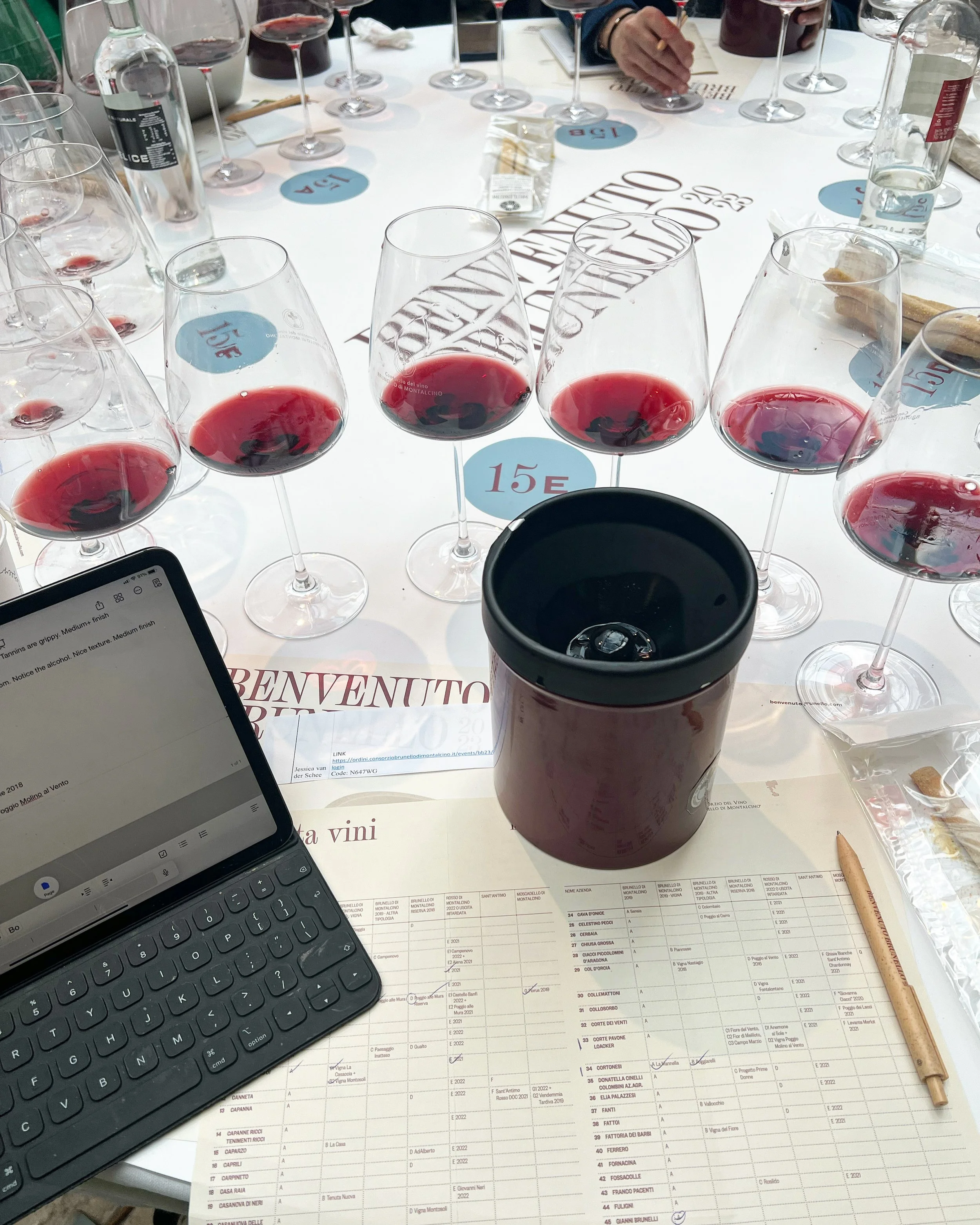 Table with multiple glasses of red wine, a wine bottle, a coffee cup, a laptop, and a sheet with wine list on it. A person's hand is holding a pen, writing on a sheet. Colored circles with numbers and prices are on the table, indicating wine varietie
