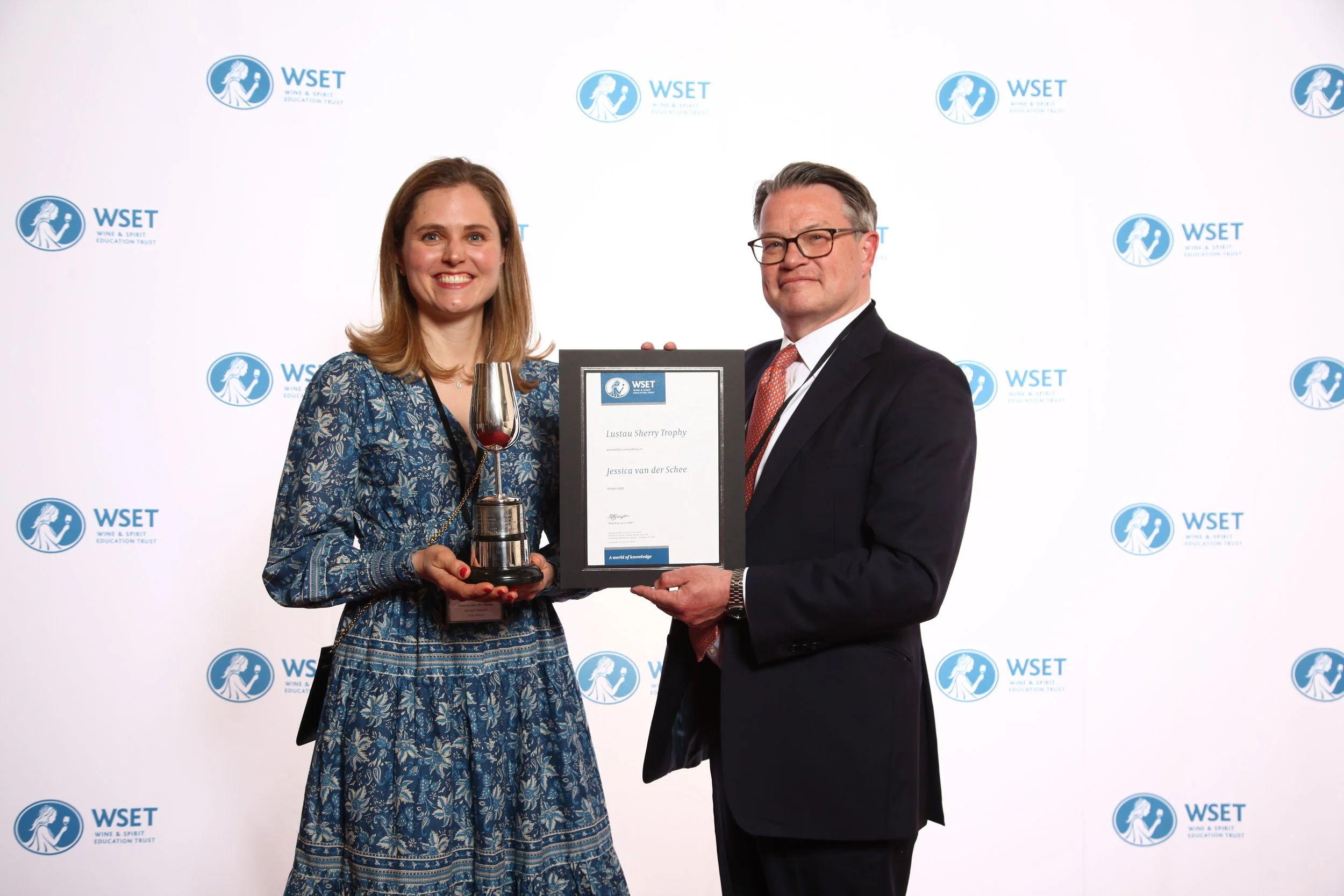 A woman and a man standing in front of a step and repeat banner with the WSET logo. The woman is holding a trophy and a glass of red wine, and the man is presenting a certificate to her.