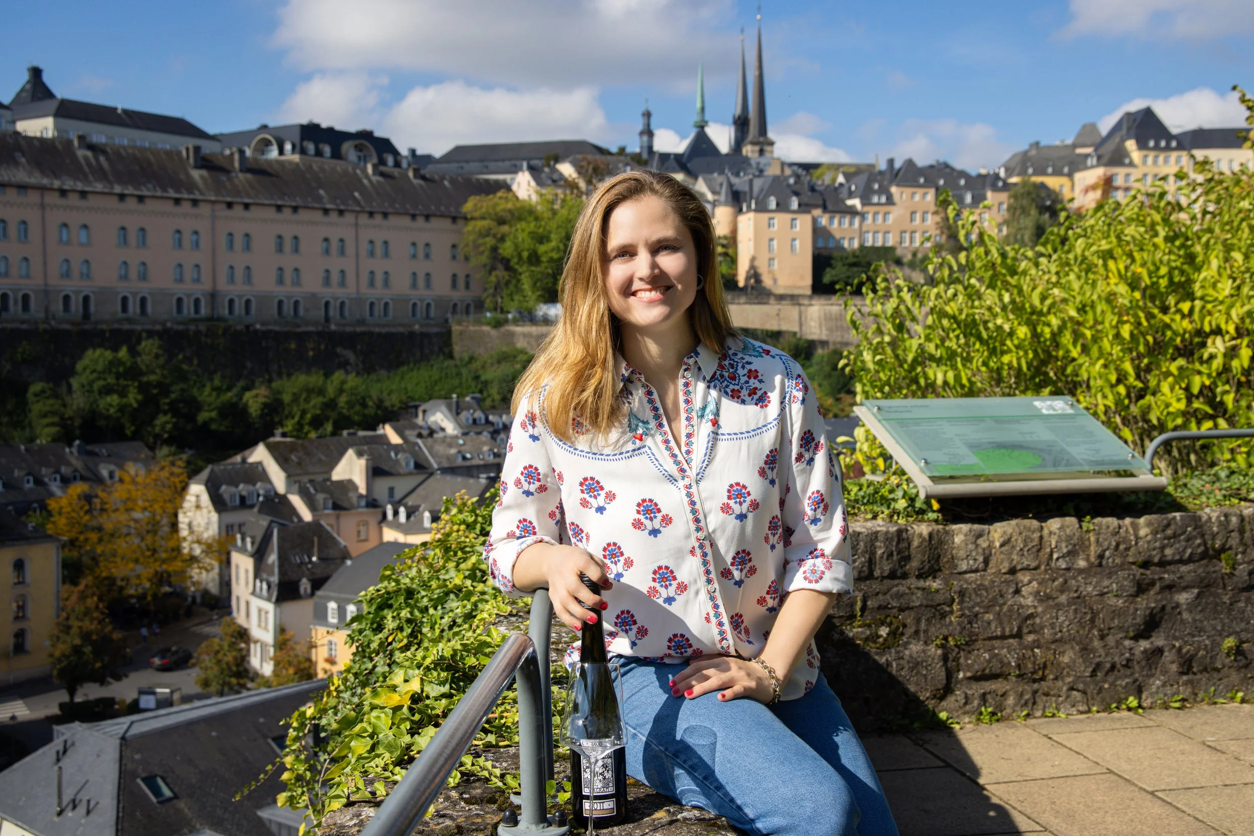 A young woman with long blonde hair smiling, sitting outdoors on a ledge with a cityscape background that includes historic buildings and church spires. She has a bottle of wine and a glass next to her.