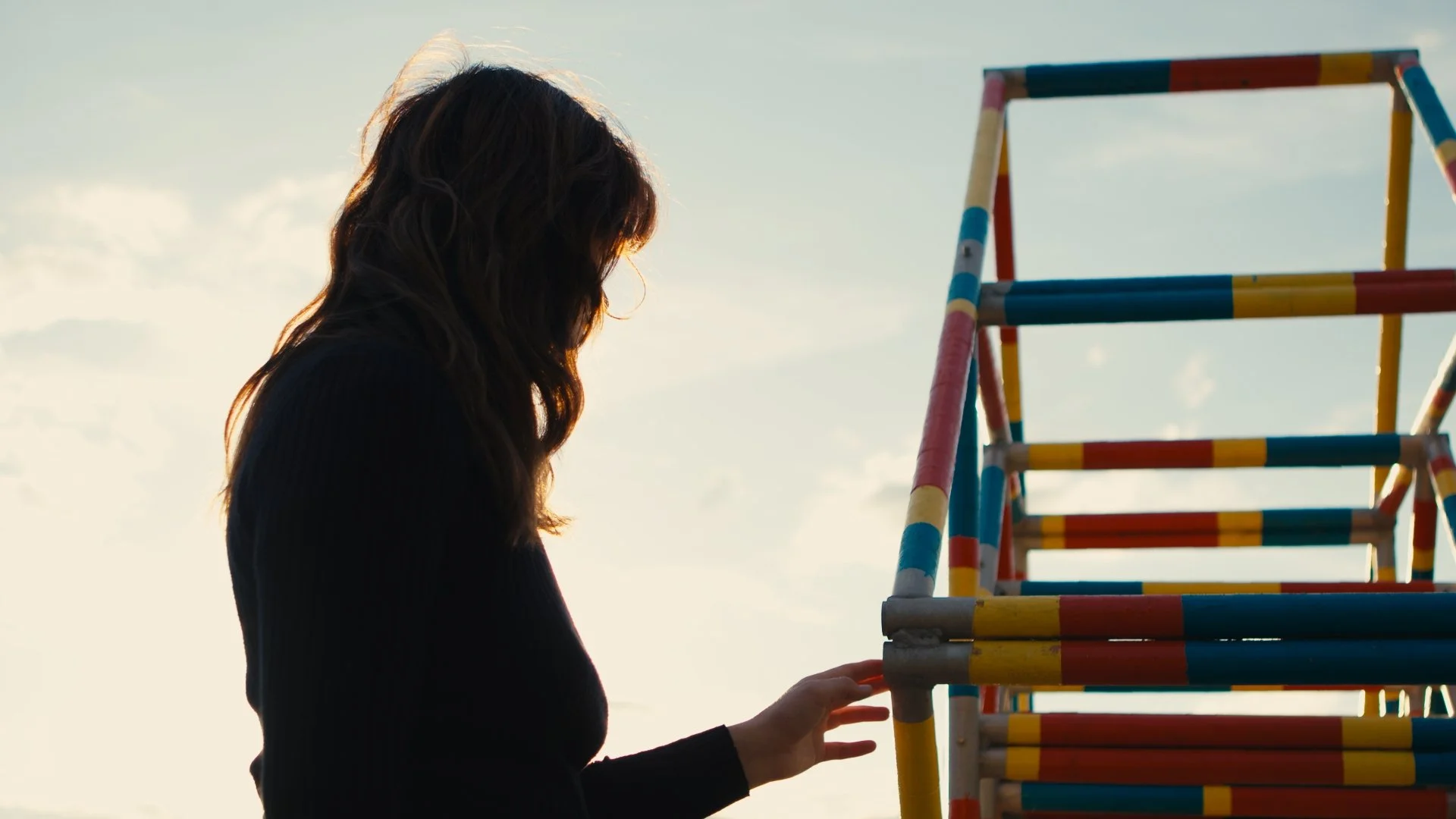 A person with brown hair and dark clothing touching a colorful abstract sculpture by Ray Arnatt outdoors against a cloudy sky.