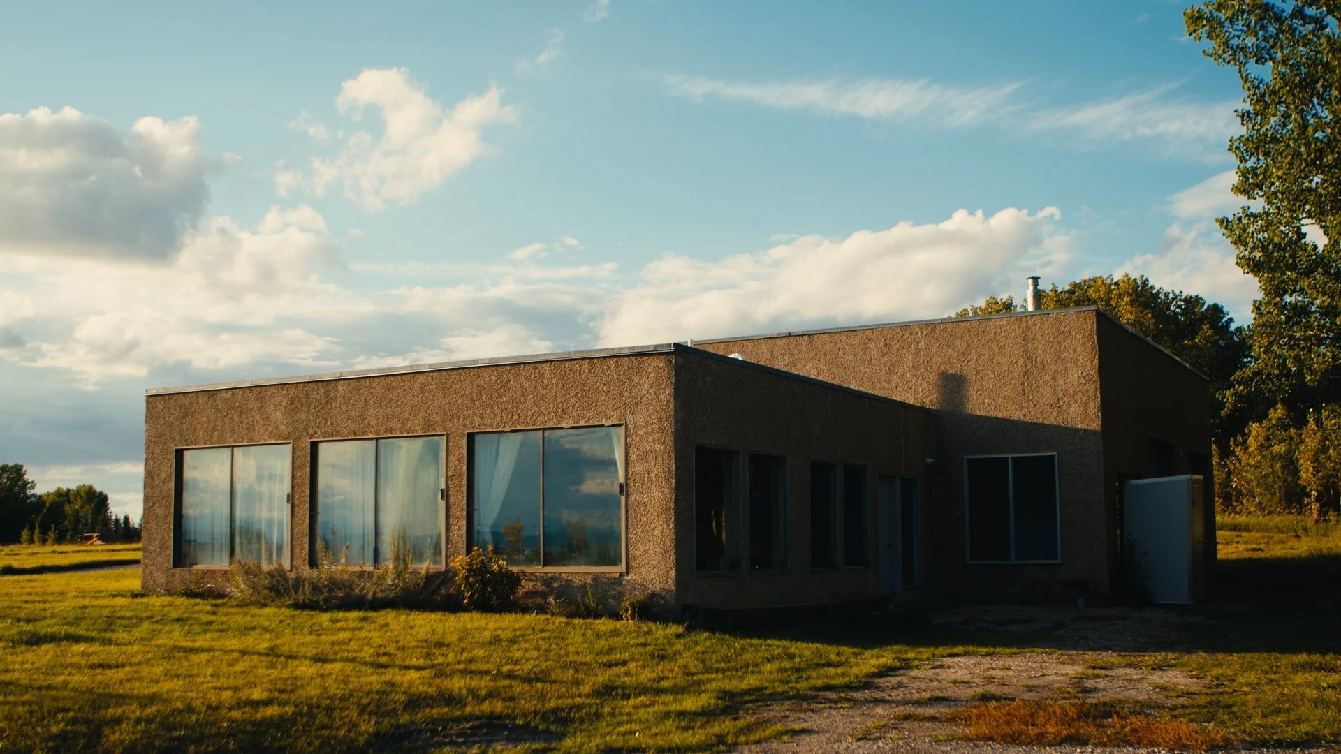 Modern studio building with large glass windows, surrounded by grass and trees, under a partly cloudy sky during sunset.