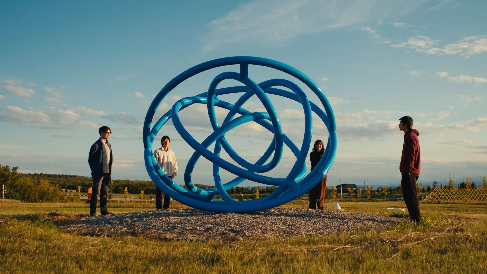 Four people standing around a large blue abstract sculpture outdoors in a field during sunset. The sculpture is 'Doodle #4' by Katie Ohe