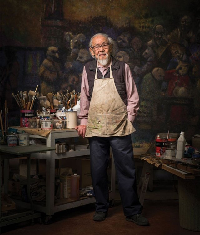Harry Kiyooka standing in his art studio surrounded by painting supplies and artworks on the wall behind him. He wears glasses, a beige apron, and a black vest over a pink shirt.