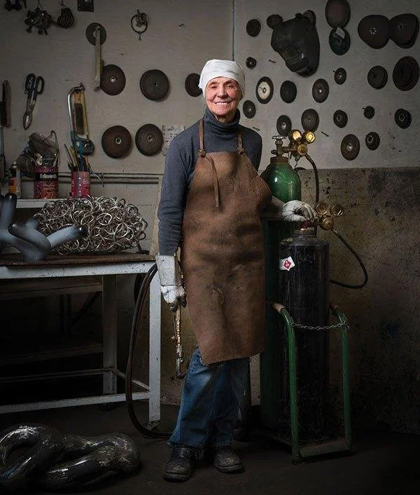 Katie Ohe wearing a white headscarf and apron standing in a welding shop with tools and metalwork on the wall.