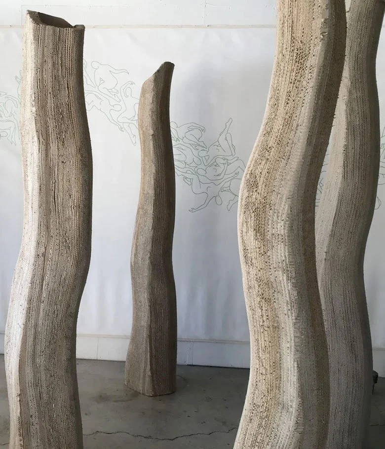 Four tall, twisted wooden sculpture pieces standing on a concrete floor against a white wall with faint line drawings.