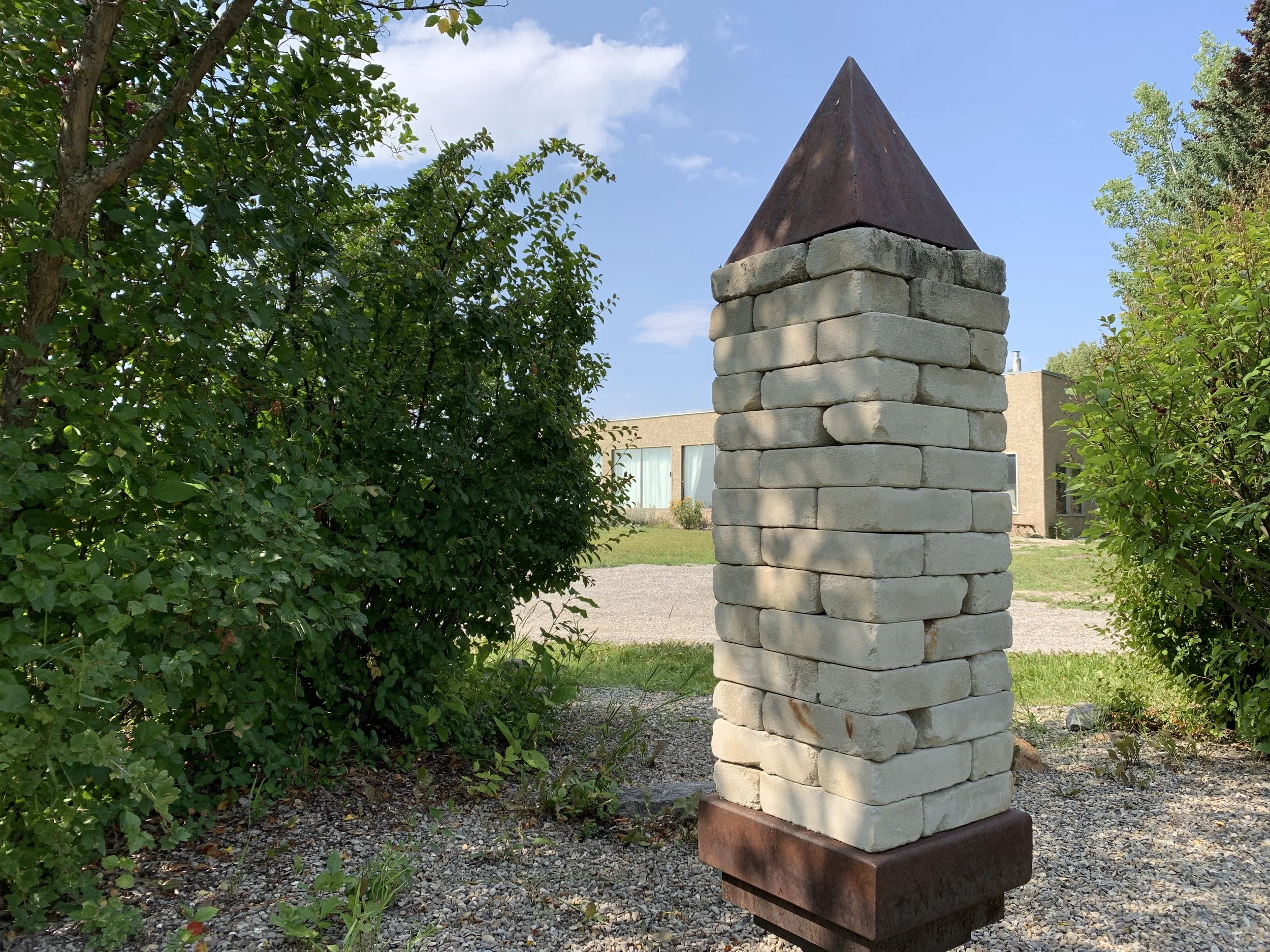 A white brick chimney sculpture by Alex Caldwell 'wheelhouse' with a pointed pyramid-like metal topper, placed outdoors on a gravel surface, surrounded by green bushes and trees under a blue sky, in the KOAC garden.