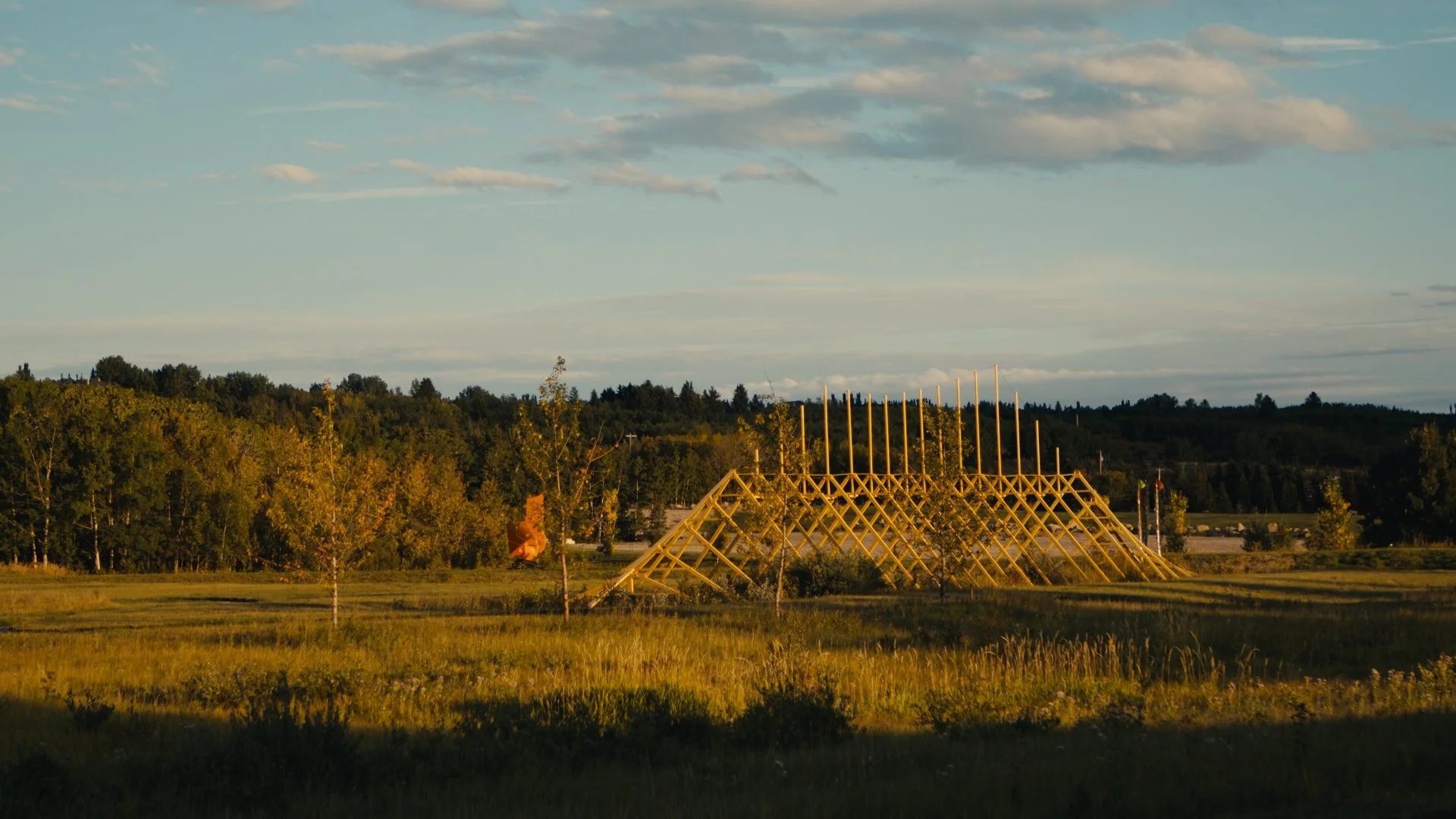 A metal sculpture 'Bridge' by Charles Boyce looks like a triangular tunnel bridge, but sits atop a grassy field with trees and hills in the background, illuminated by warm sunlight. The sculpture is painted bright yellow.