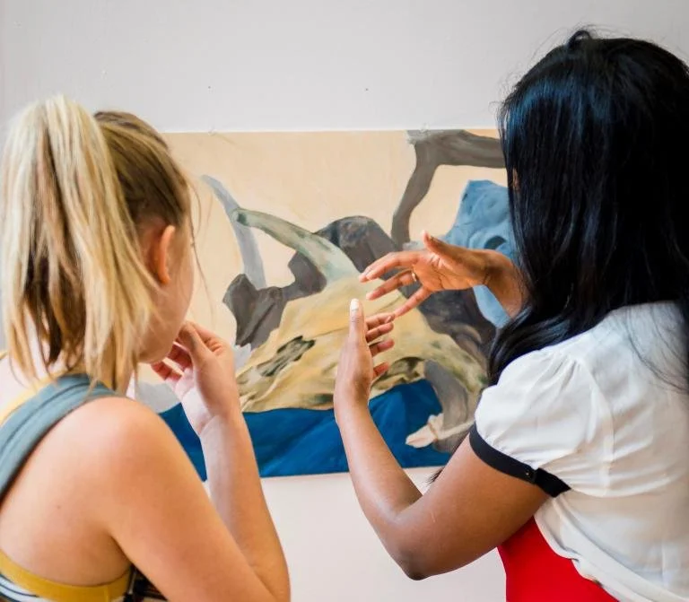 Two women discuss a painting of a landscape with mountains and trees; one has blonde hair and the other has black hair with blue highlights.