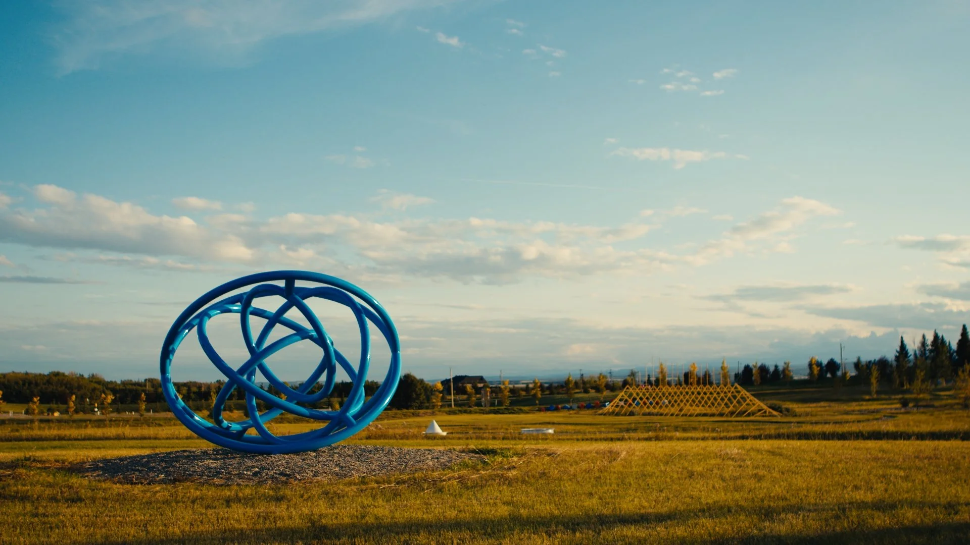 An outdoor park scene with a large blue abstract spherical sculpture 'Doodle #4' by Katie Ohe in the foreground, grassy fields, a yellow geometric structure in the distance, and a blue sky with scattered clouds.