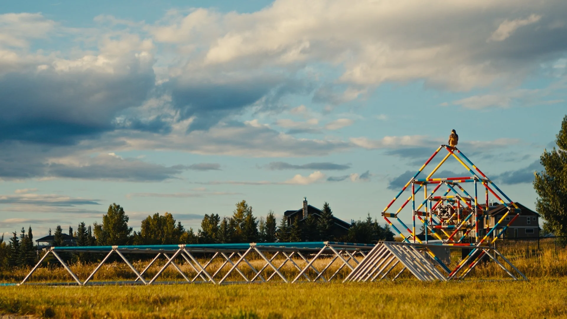 An abstract sculpture 'Binder' by artist Ray Arnatt (which resembles a jungle gym with steel bars painted in bright red, yellow, and blue) is sitting in a grassy field under a partly cloudy sky, with houses and trees in the background.