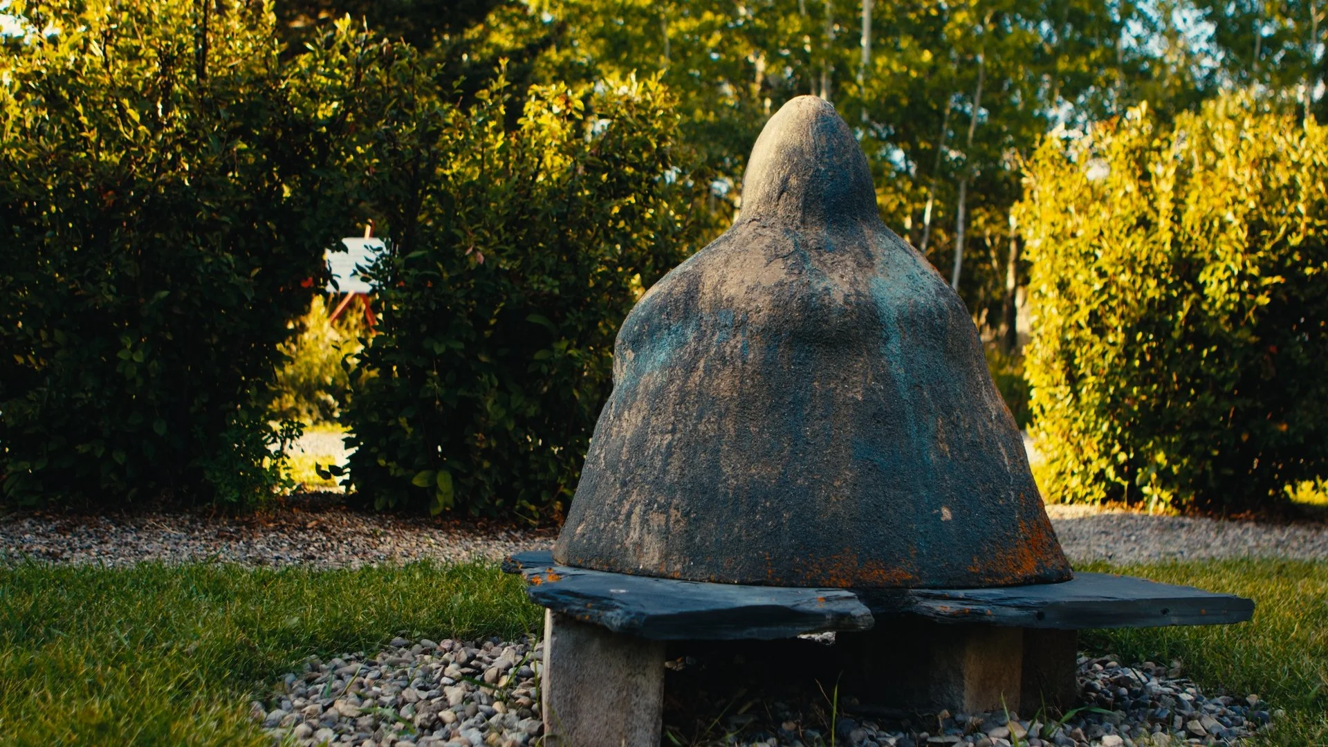 Stone sculpture 'Earth Mother' by Katie Ohe resembling a non-descript figure on a wooden platform in the KOAC outdoor garden, surrounded by grass, bushes, and trees in the background.
