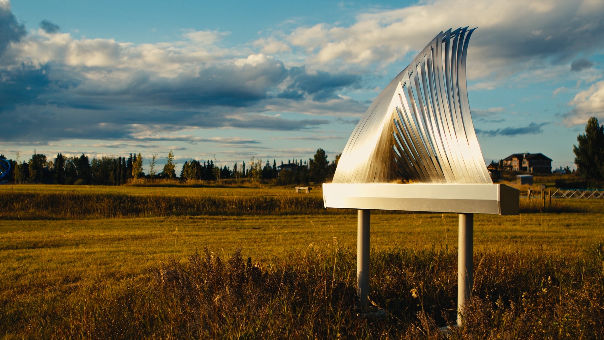 A metallic outdoor sculpture with curved, slatted design in a grassy field under a cloudy sky. The sculpture is titled 'Shimmering Willow' by James Ziegler.