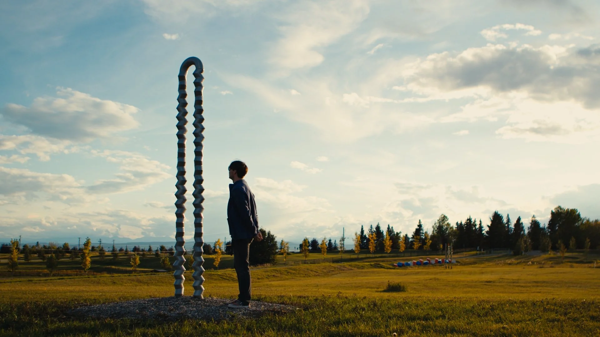 A figure stands outdoors in the sculpture park at sunset, looking at a tall, twisted sculpture made of interconnected segments - ZigZag by Katie ohe - with trees and a cloudy sky in the background.