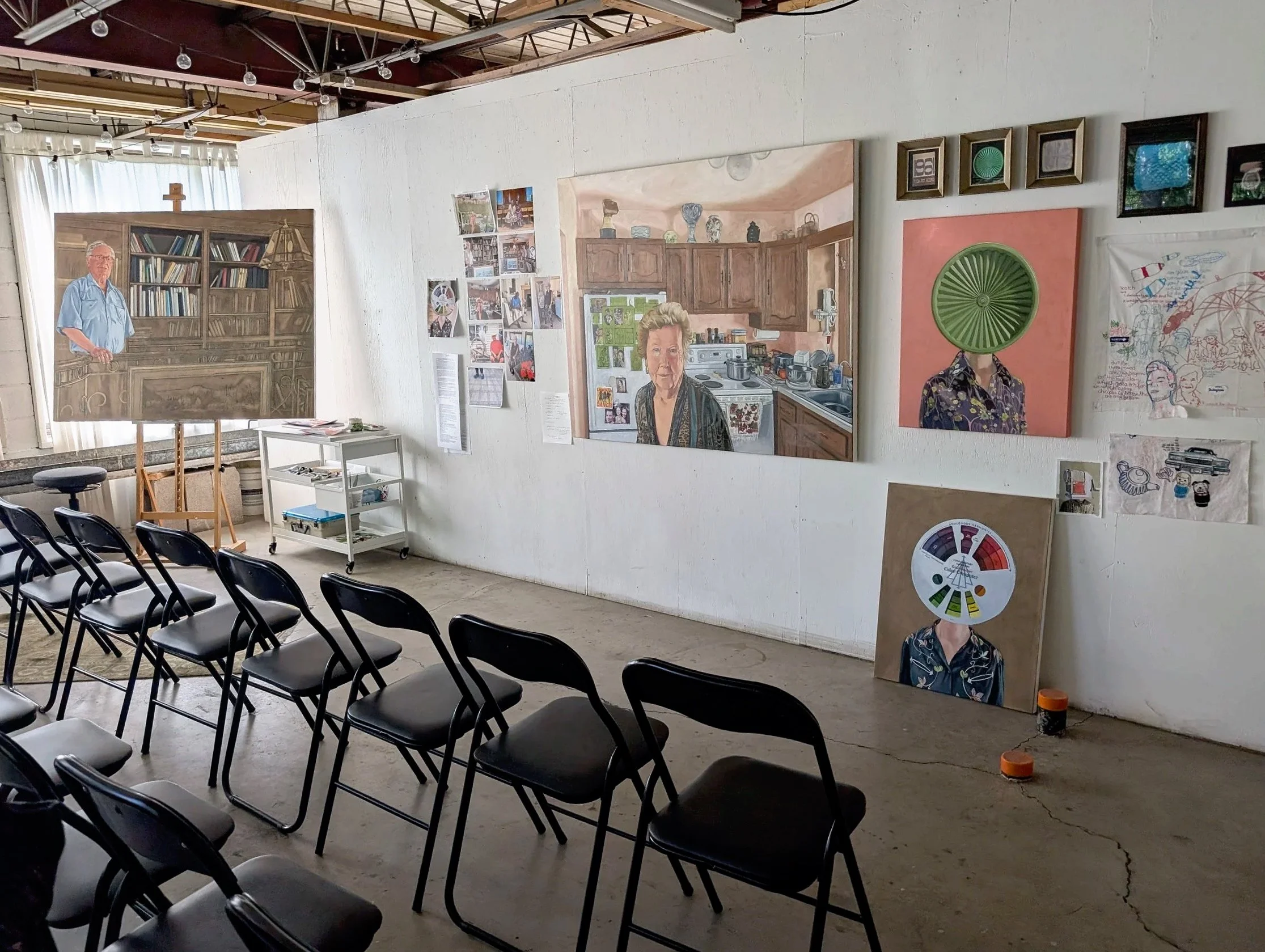 The KOAC residency studio with work by Pamela Vickerson featuring multiple portraits and mixed media art pieces on a white wall, with black folding chairs arranged in front, and a white rolling cart below the artwork.