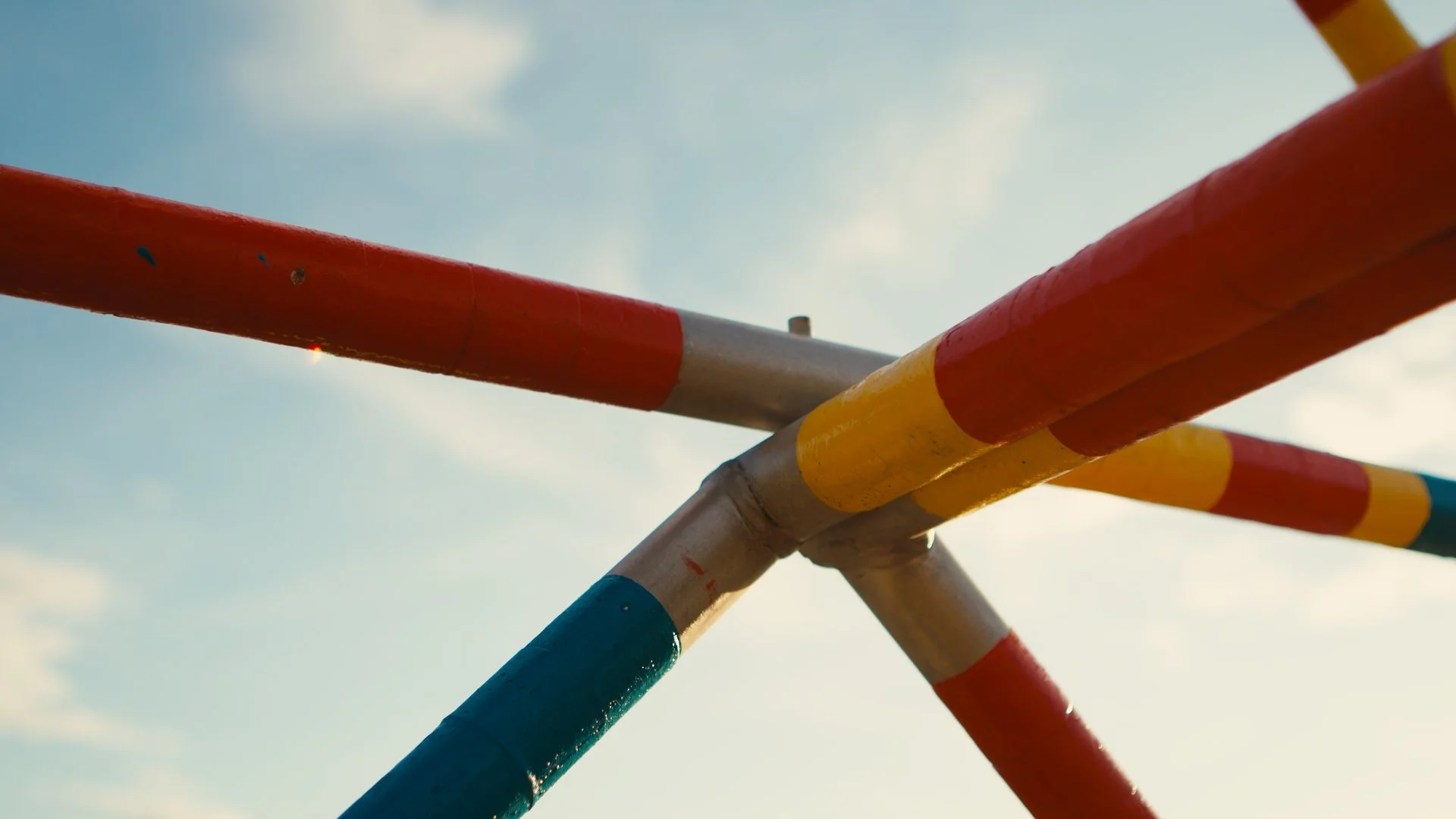 Coloured sculpture detail - that looks like a playground structure - with blue sky background. The sculpture is by artist Ray Arnatt and is titled 'Binder' - it is structured like a jungle-gym with steel bars painted in bright red, blue, and yellow.