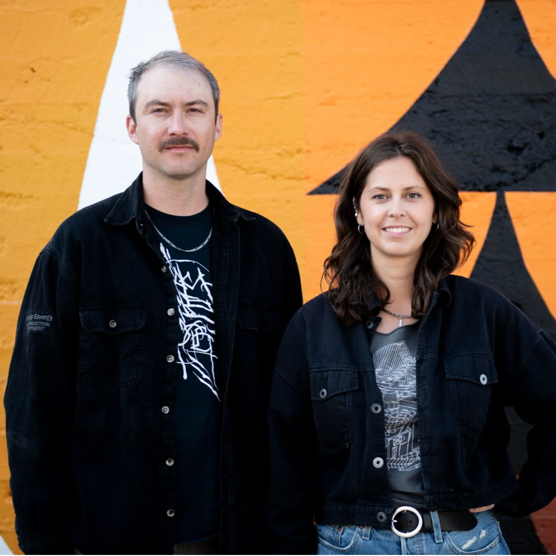 An image of the artist duo NASARIMBA. Mikhail Miller and Rachel Ziriadaare shown in front of one of their colourful murals with orange, black, and white geometric sections.