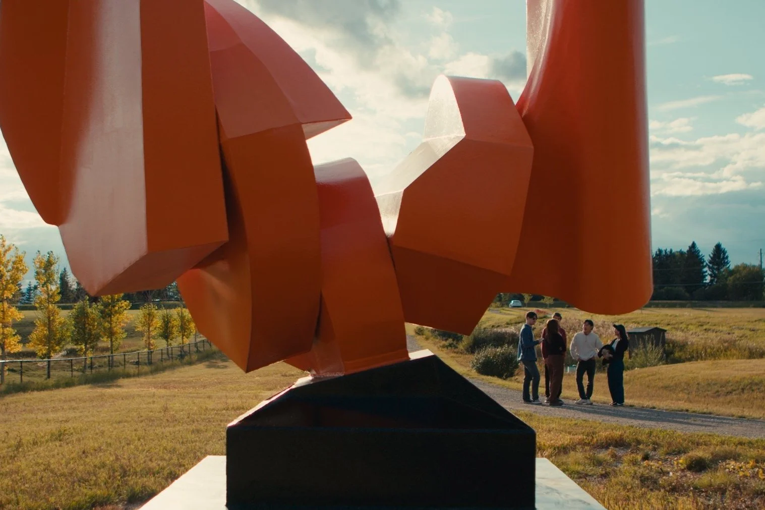 A large abstract sculpture 'Steel Wave' by Roy Leadbeater with bold orange geometric shapes in a park setting during daytime, with four people standing and talking in the background and trees with fall foliage.
