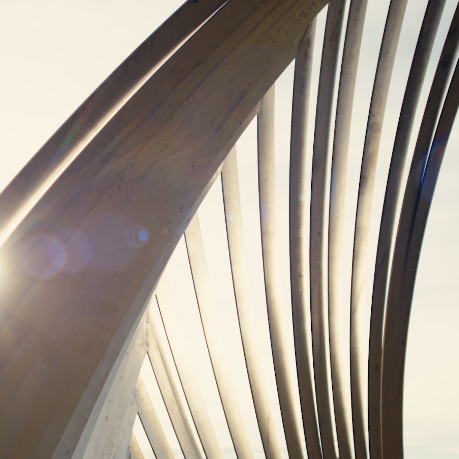 Close-up of a modern architectural sculpture with curved metal beams and supports, where sunlight is creating a lens flare. The sculpture is by artist James Ziegler titled 'Shimmering Willow'
