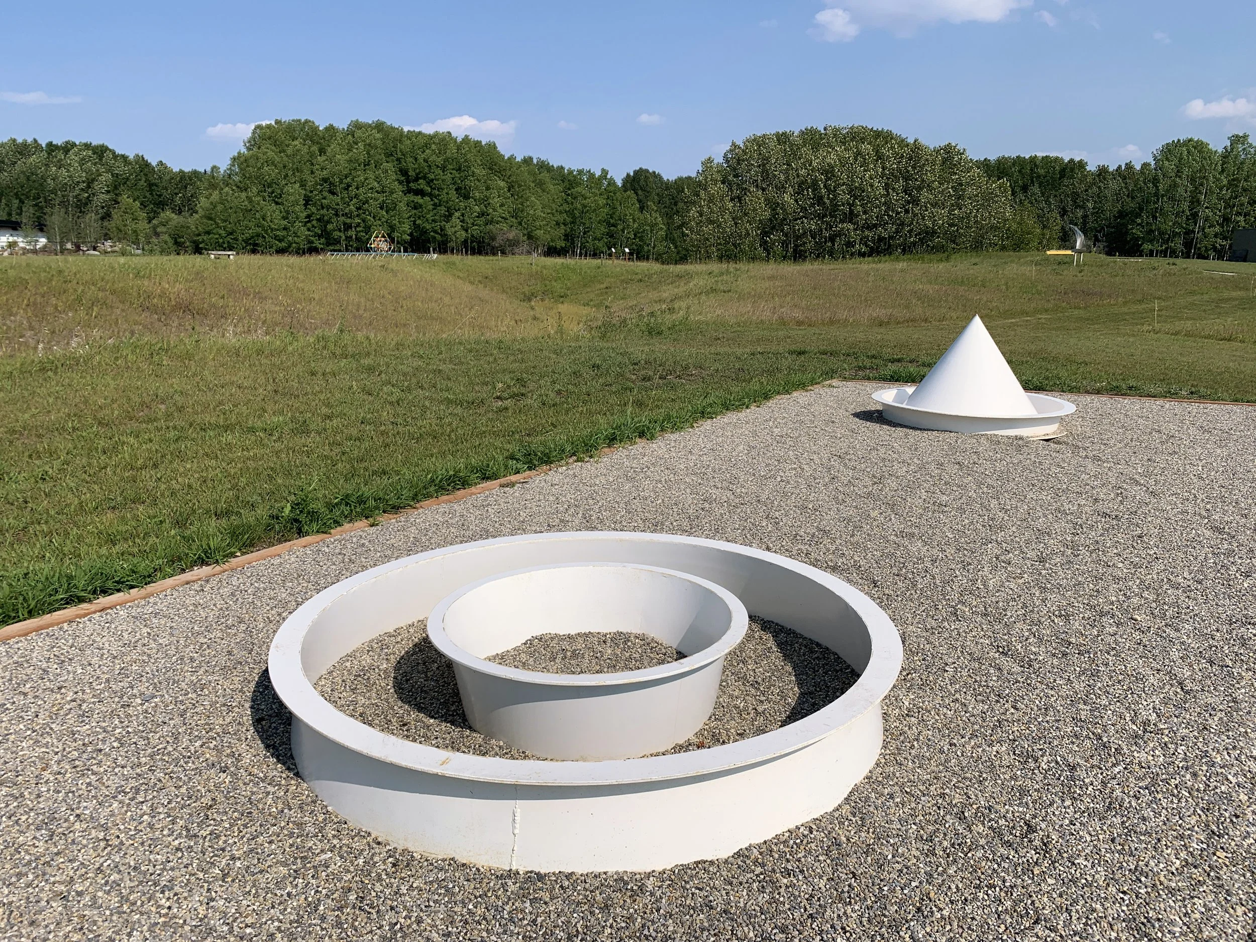 'Kiyooka Garden' is a sculpture by Katie Ohe with white geometric shapes on gravel ground, including a cone and a circular structure, in a grassy park with trees and blue sky in the background.