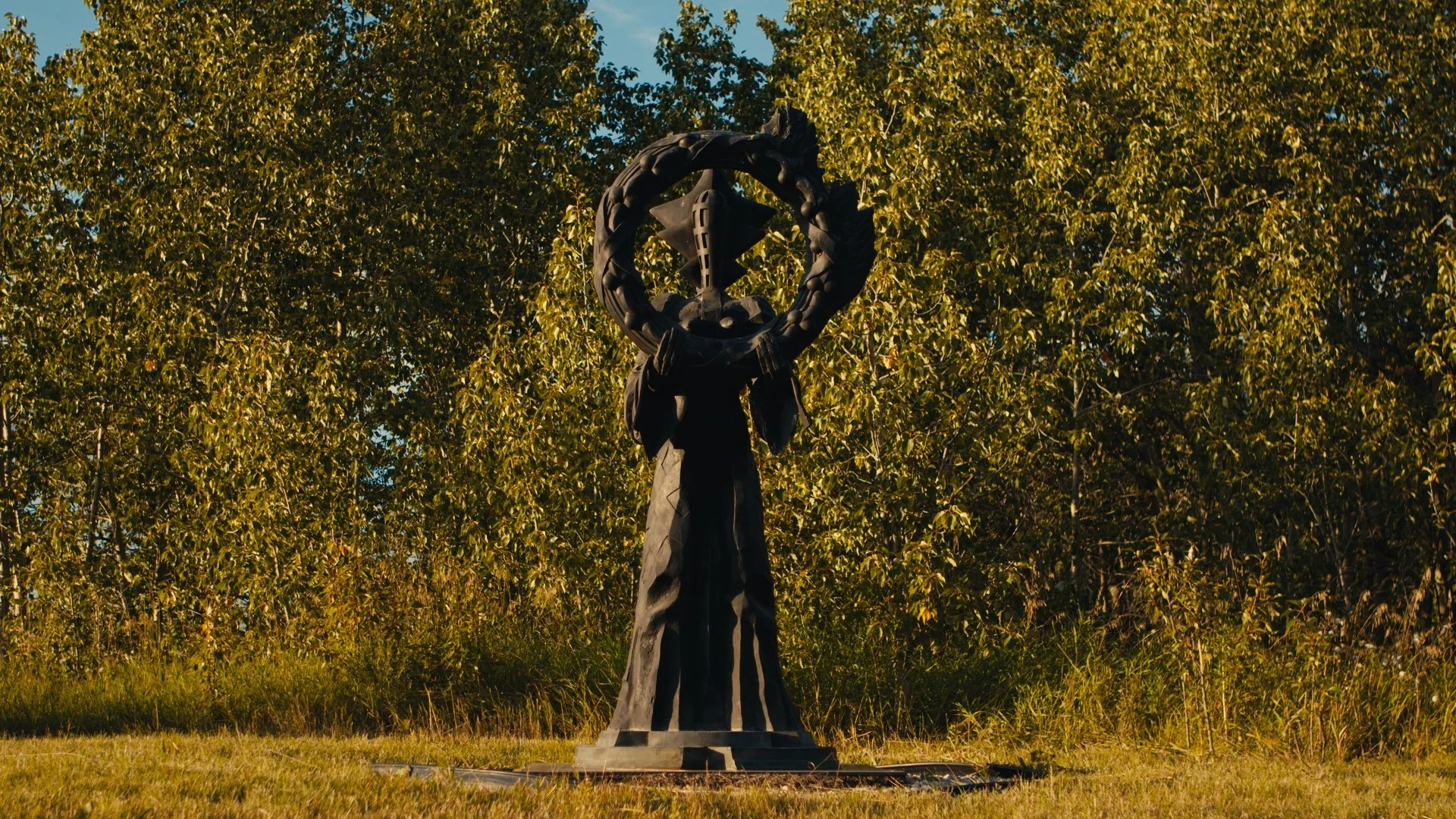 A dark metal sculpture by artist Michael Sandle 'Queen of the Night'. The sculpture is a figure holding a large circular wreath, in a grassy area with trees in the background.