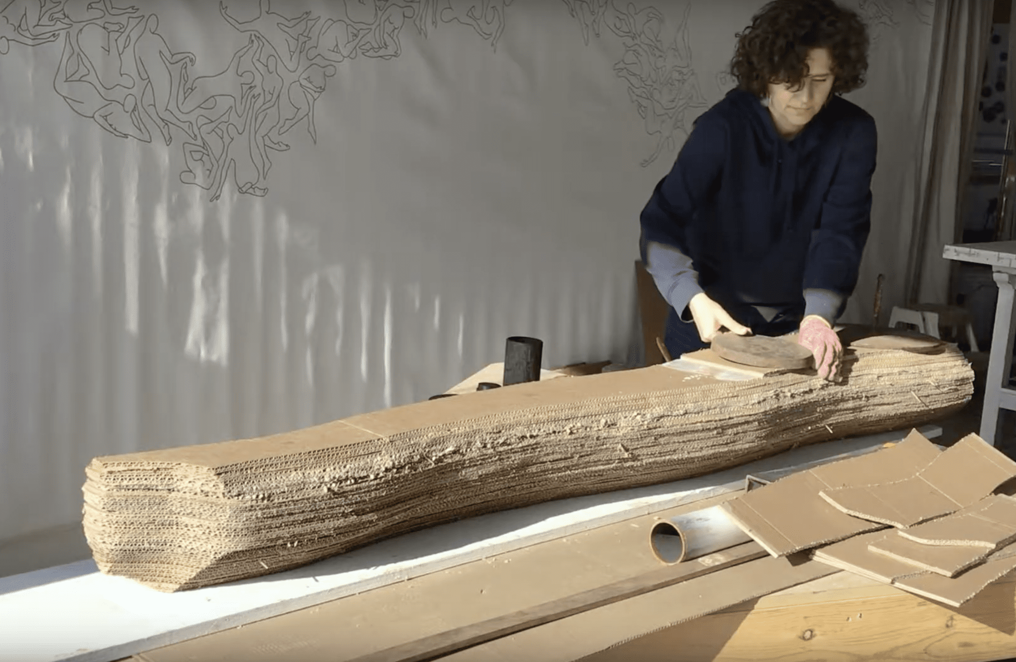Artist Patricia Lortie wearing a dark hoodie and working on a large piece of layered cardboard, using a cutting tool, in a studio with a white background and an abstract line art on the wall.