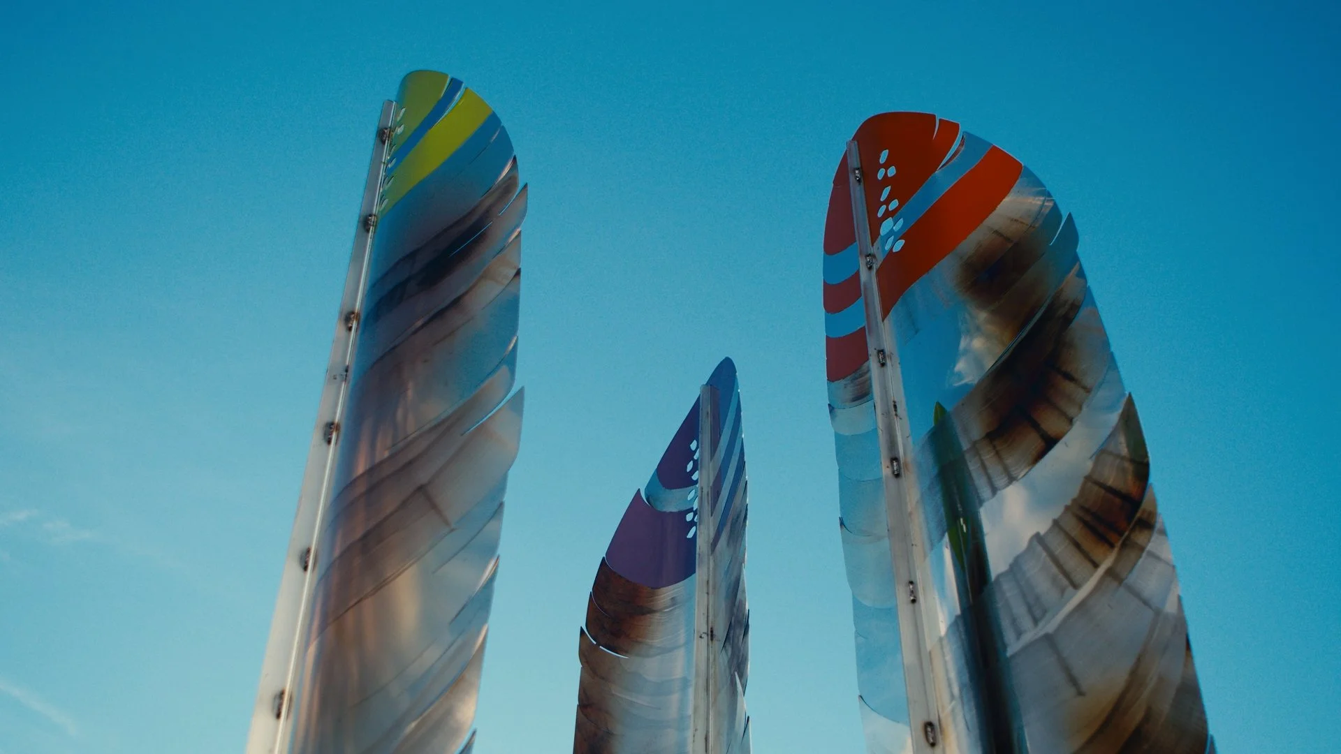 Three tall, reflective metallic sculptures resembling feathers or leaves against a clear blue sky. This sculpture is by James Ziegler 'Tall Feathers' and was developed through collaboration with Blackfoot Elder Harley Crowshoe.