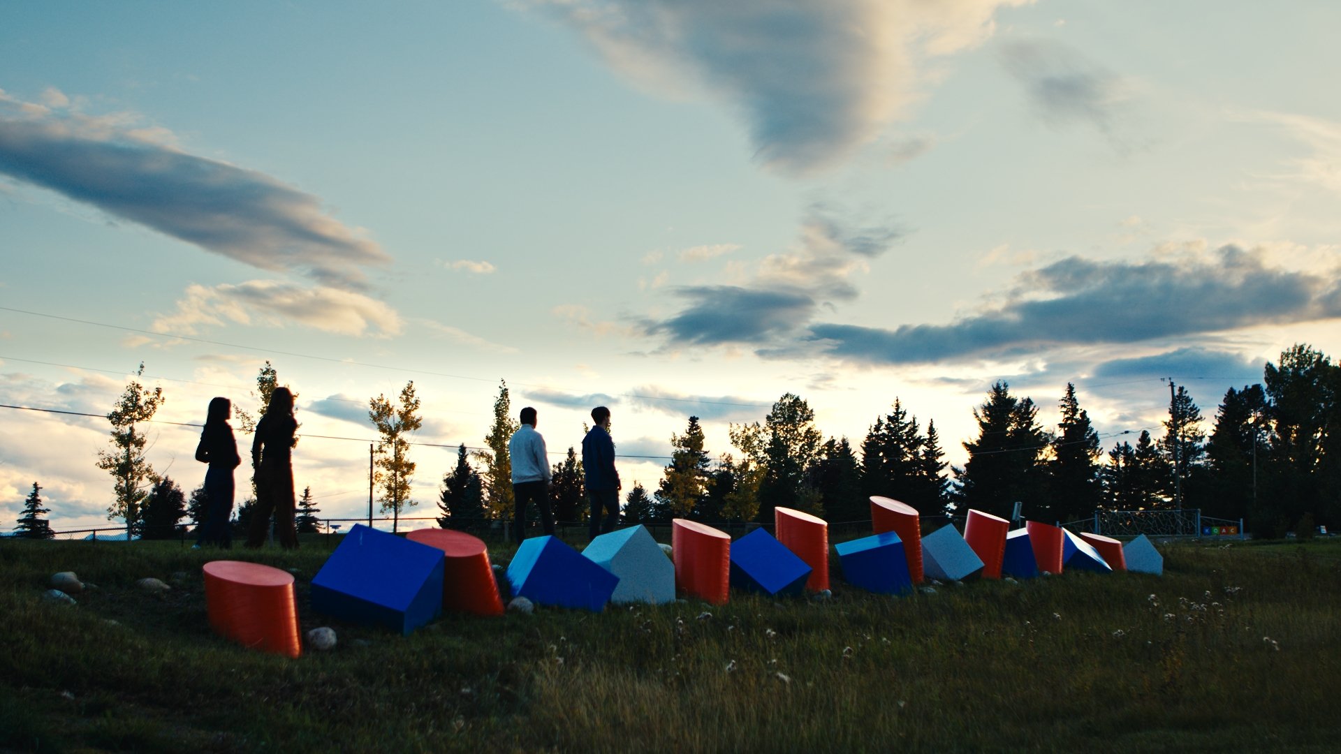 Four people silhouetted against a sunset sky walk past colourful geometric block sculptures on a grassy hill, with trees and clouds in the background. The sculpture grouping is named 'Blocks' by artist Ray Arnatt - with blocks in blue, red and white