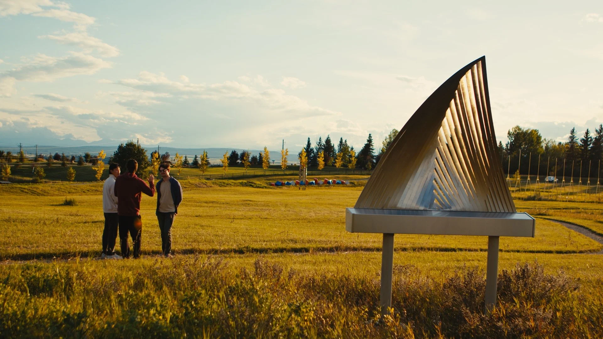 Three people standing in a grassy field with a large, modern metal sculpture by James Ziegler in the foreground and trees in the background.