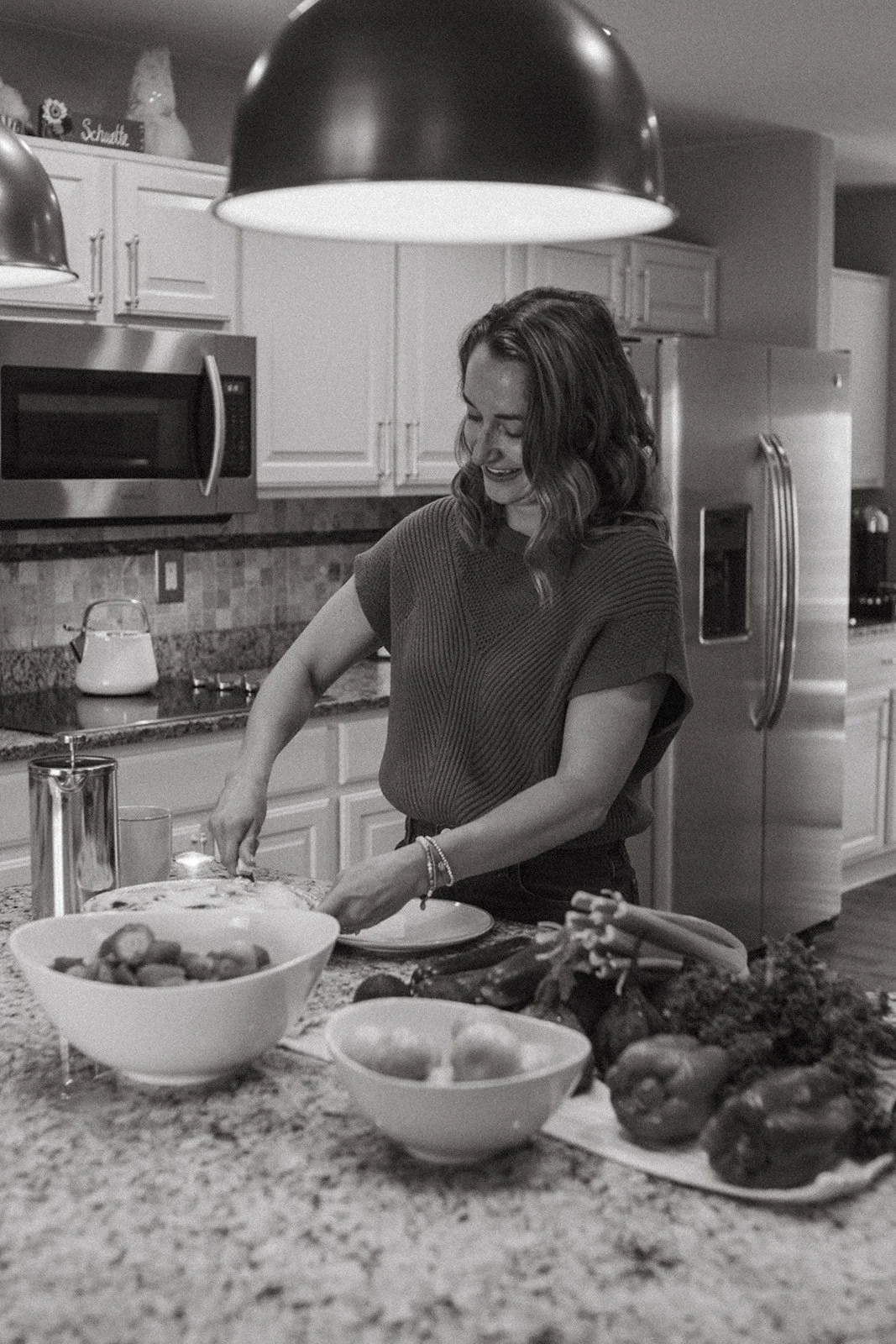 A woman cooking in a kitchen, preparing food on a granite countertop. There are vegetables and bowls on the counter, with a refrigerator and microwave in the background. The lighting is soft and the image is in black and white.