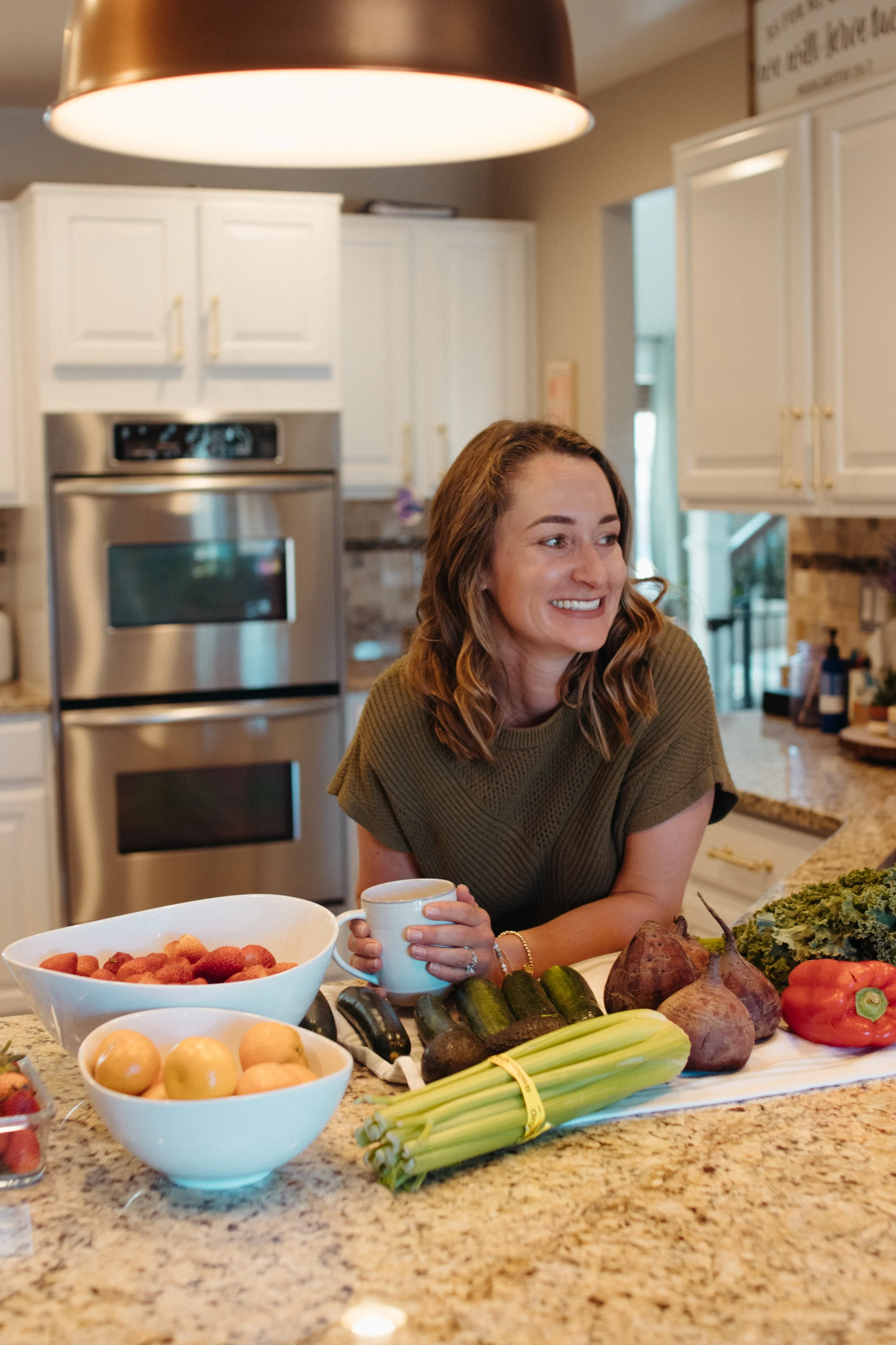 Woman in a kitchen with assorted fresh produce, including strawberries, grapefruits, celery, zucchini, sweet potatoes, kale, and a red bell pepper, smiling and holding a mug.