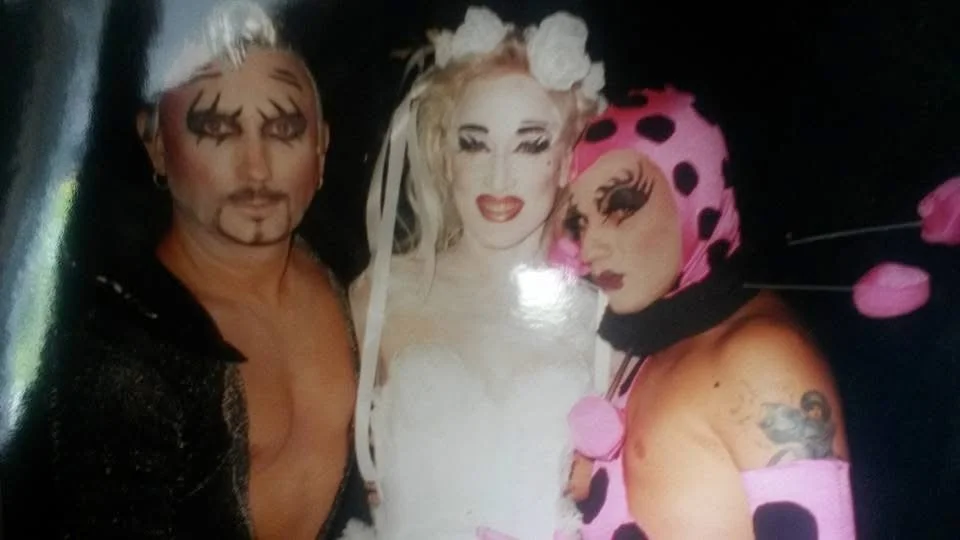 Three people with elaborate makeup and costumes, posing together against a dark background.