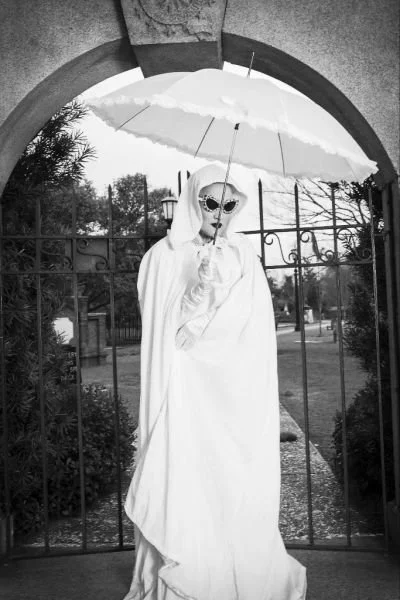 A person dressed in a white cloak and sunglasses holds an umbrella while standing outdoors behind a gate.